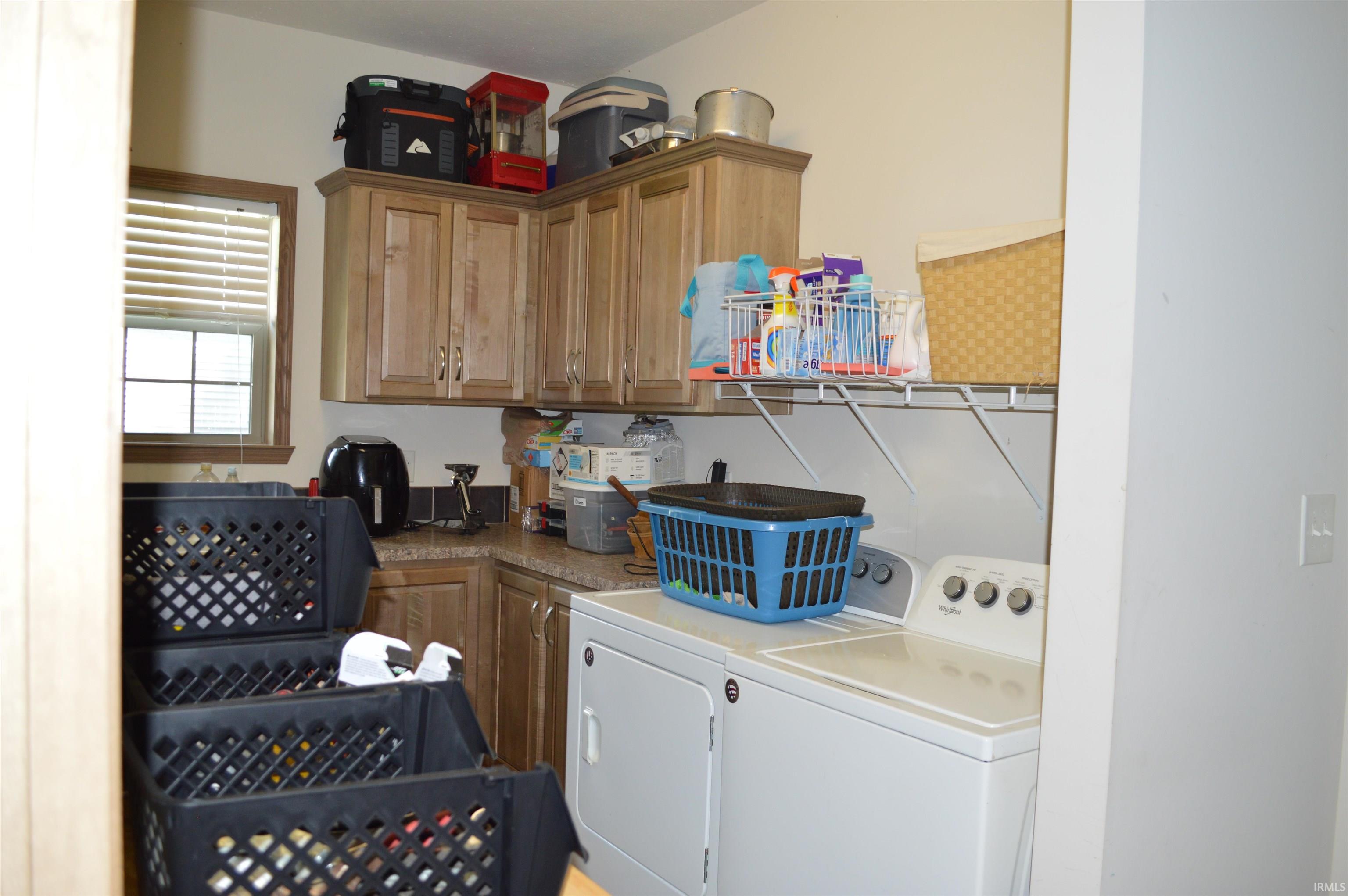 Dedicated utility space featuring light wood-finish cabinetry, a window with horizontal blinds, a countertop, and a wall-mounted shelving unit