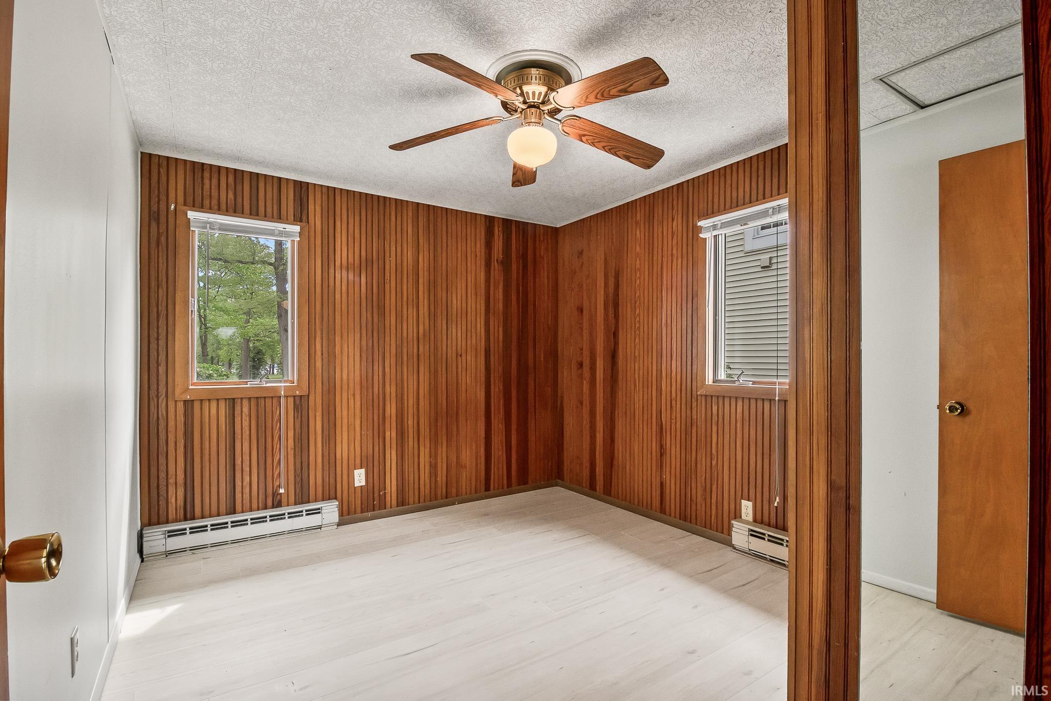 Interior room featuring wood paneling, light wood-finish flooring, two windows with blinds, and a ceiling fan with integrated lighting