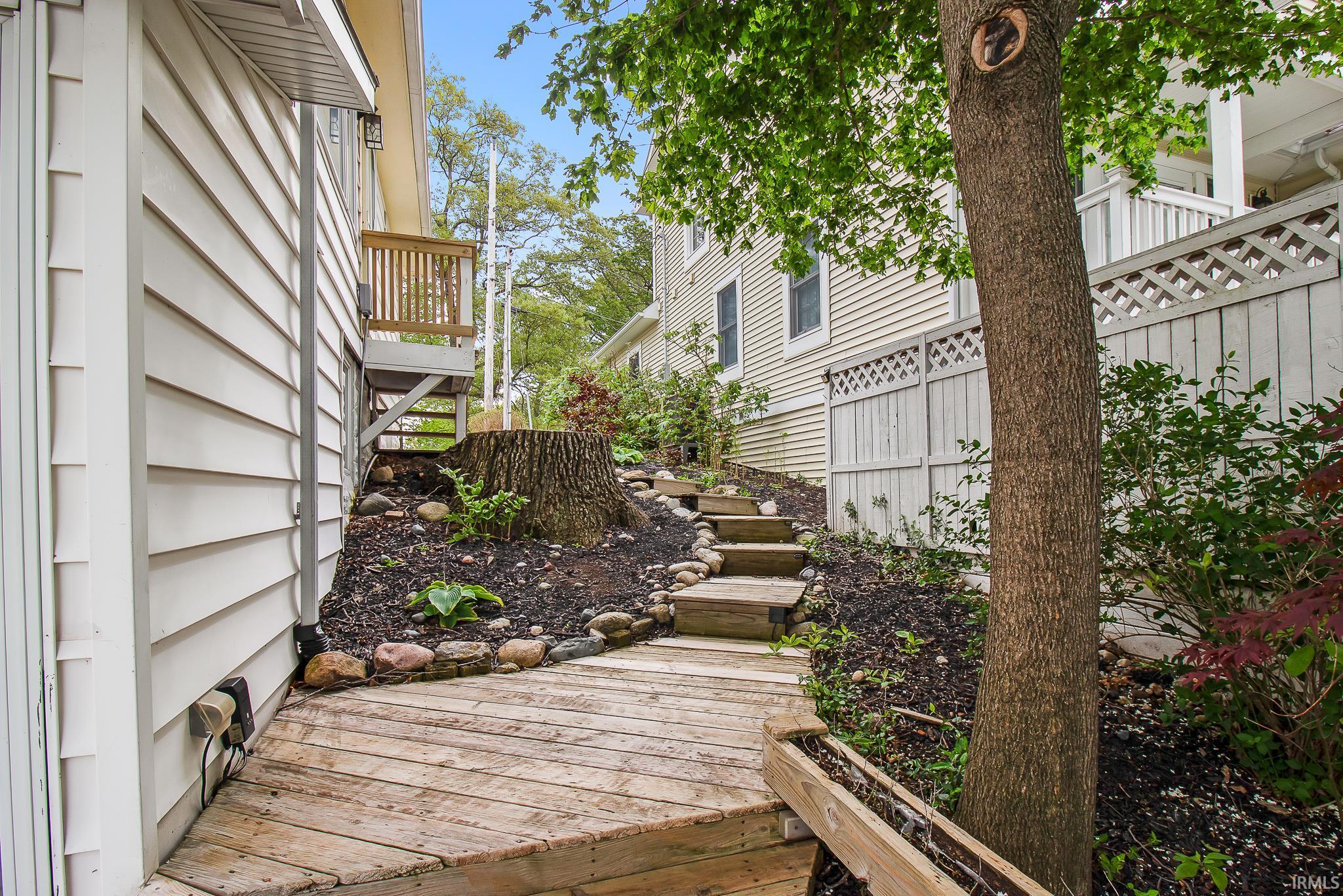 Exterior wood-finish ramp leading to a terraced garden with wood steps, featuring a mature tree and established plantings