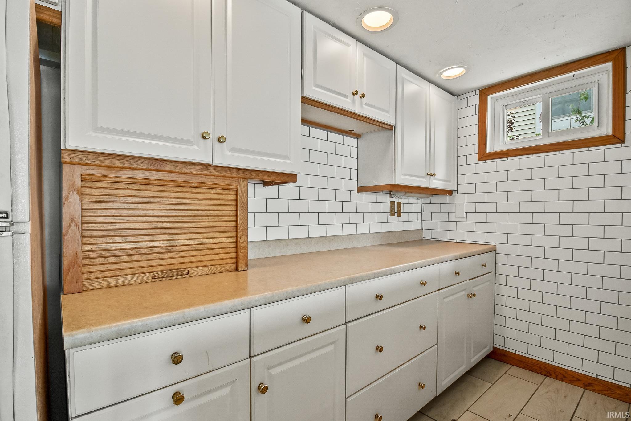 Kitchenette featuring white cabinetry with brass hardware, light-toned countertops, subway tile backsplash, recessed lighting, and a wood-framed window