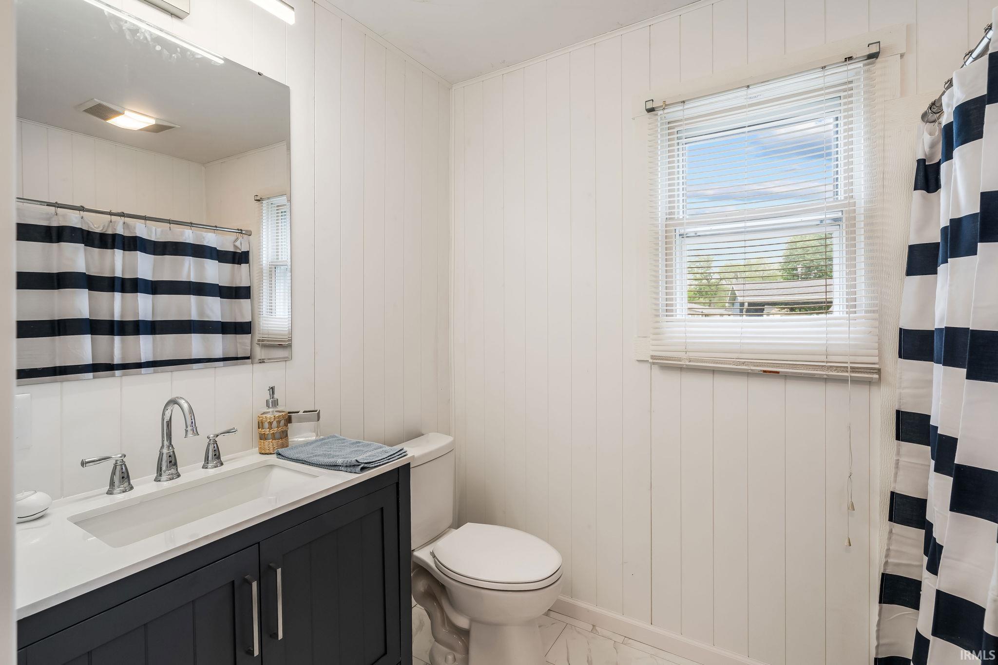 Bathroom featuring a dark wood vanity with a white countertop and integrated sink, chrome faucet, wall-mounted mirror, and a window with blinds