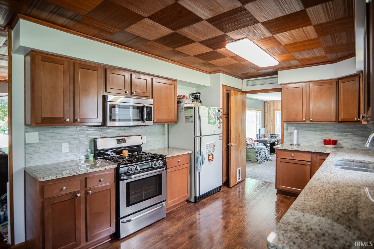 Kitchen featuring a checkered wood-finish ceiling, wood-finish flooring, brown cabinetry, light-toned granite countertops, and stainless steel appliances