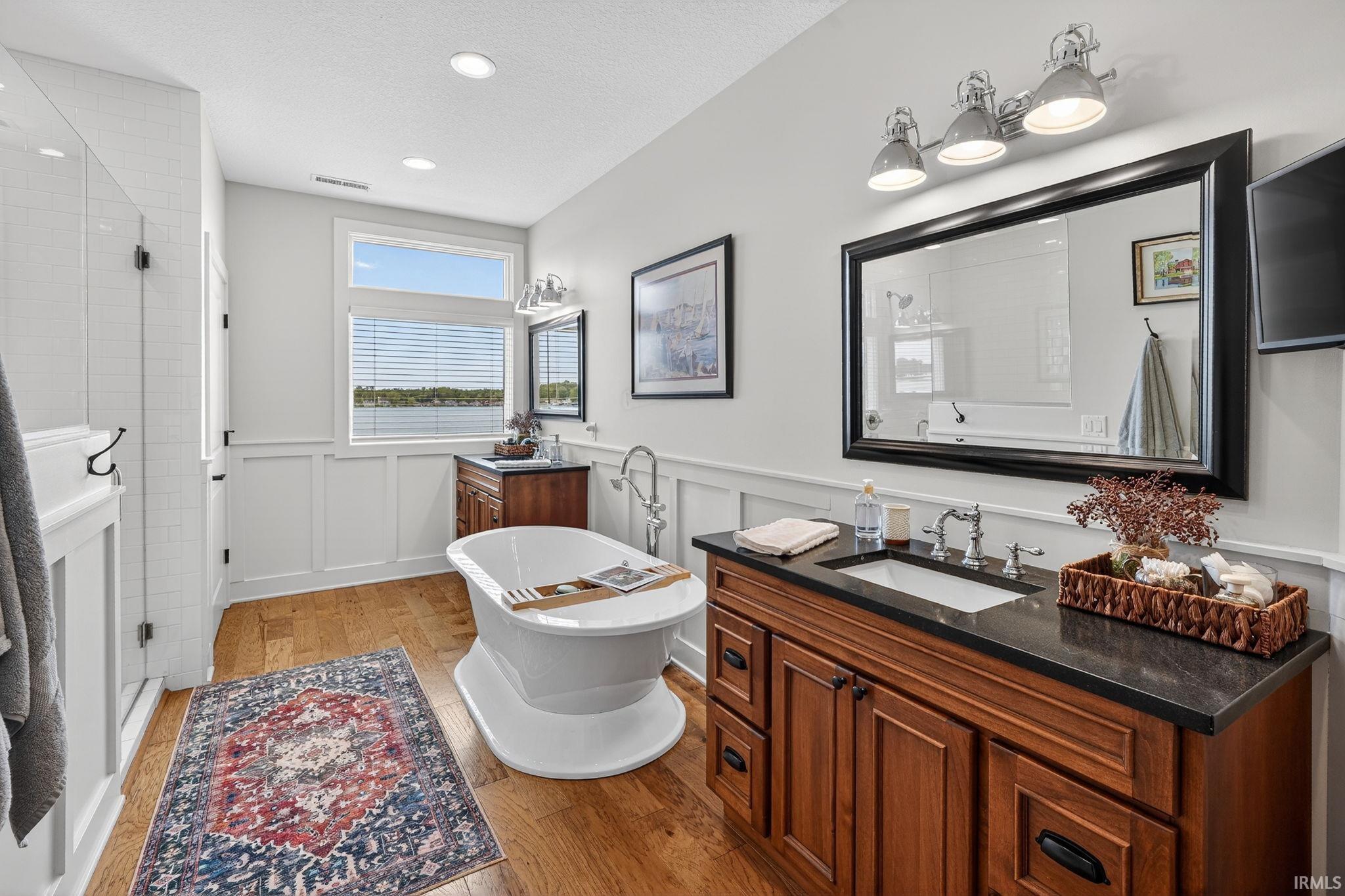 Bathroom featuring hardwood flooring, wainscoting, a freestanding soaking tub, a dark wood-finish vanity with an undermount sink, and a glass-enclosed shower