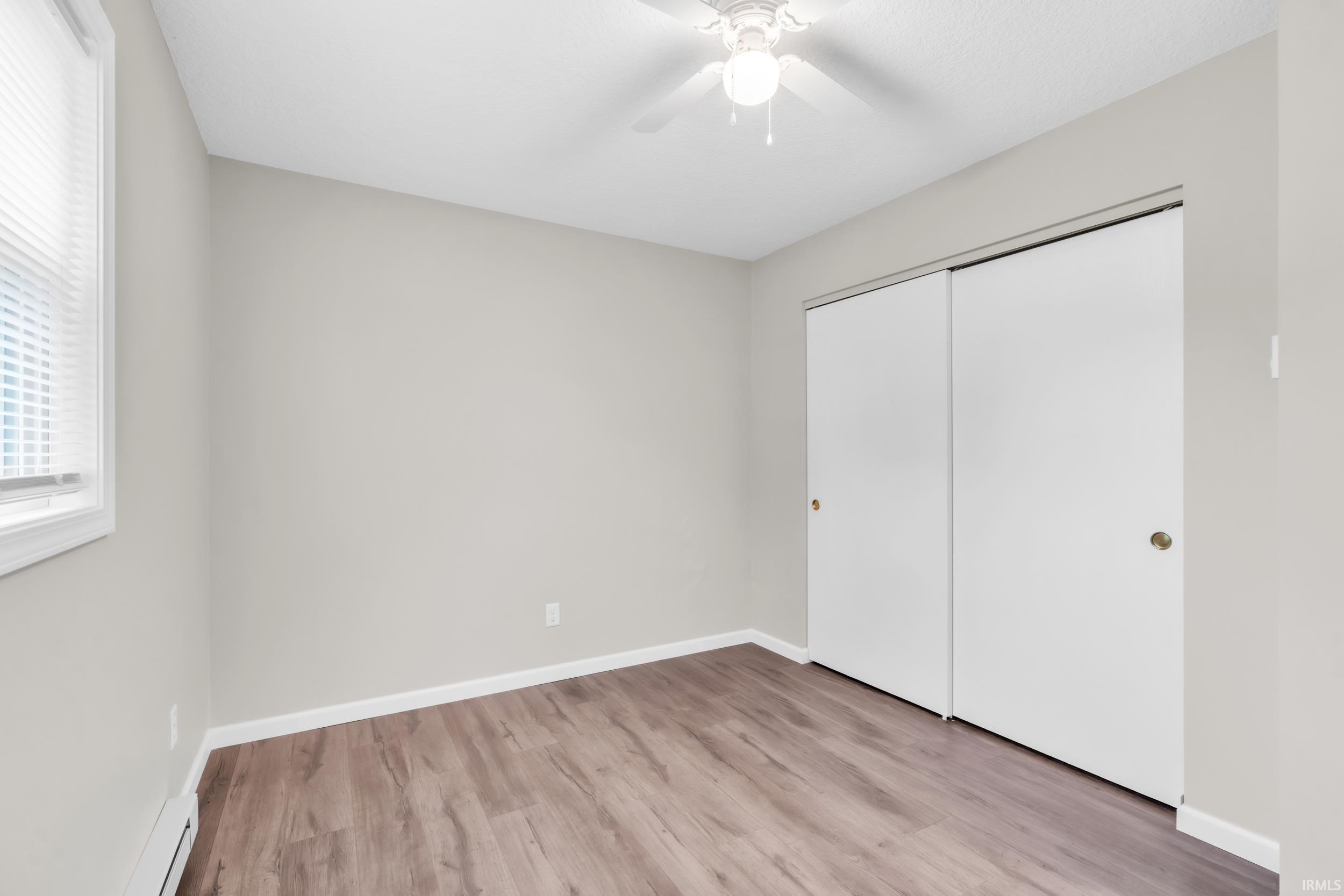 Neutral-toned room featuring wood-finish flooring, a window with blinds, a ceiling fan with integrated lighting, and white sliding closet doors