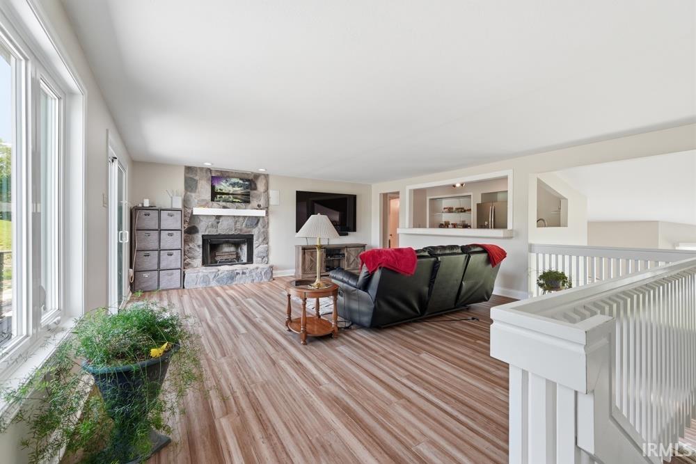 Spacious living area featuring wood-finish flooring, a stone fireplace with a substantial mantle, and ample natural light from large windows