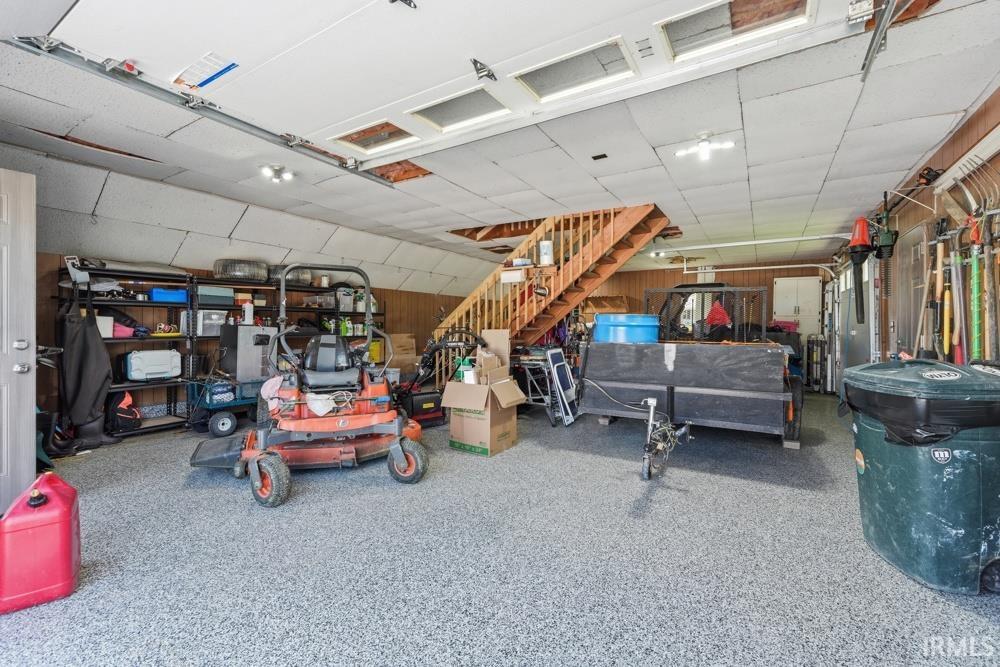 Spacious garage featuring a durable epoxy floor, overhead lighting, wood paneling, an interior staircase, and dual overhead garage doors
