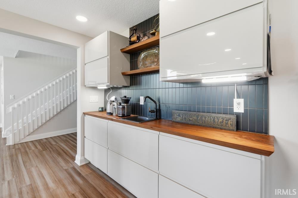 Modern kitchen area featuring light wood-finish flooring, white cabinetry, a dark tile backsplash, and wood countertops