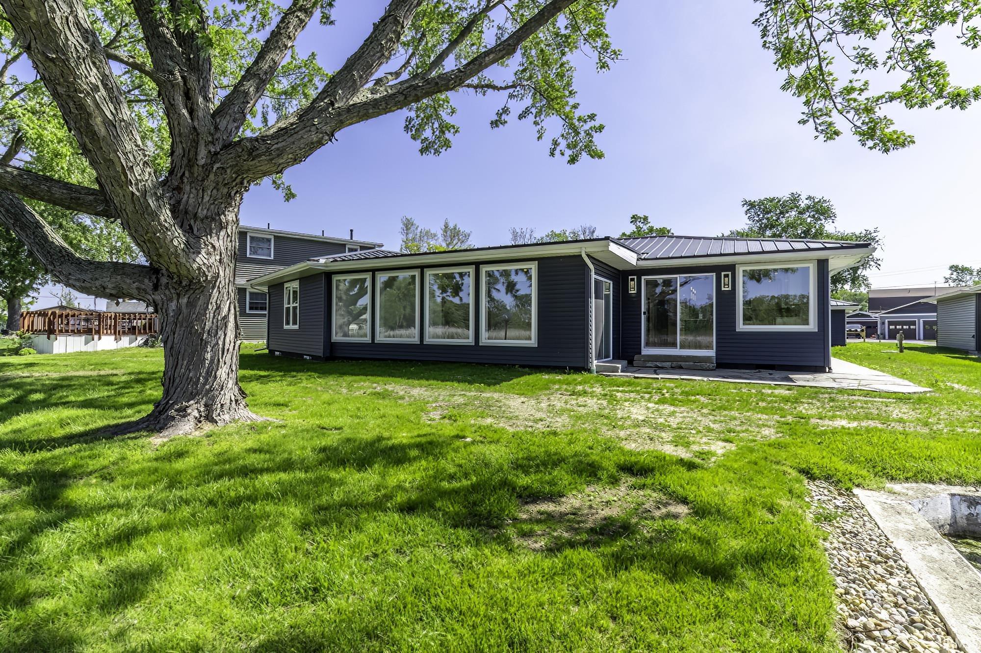 Contemporary siding exterior with a metal roof, expansive windows, a sliding glass door, an outdoor patio area, and a large mature tree