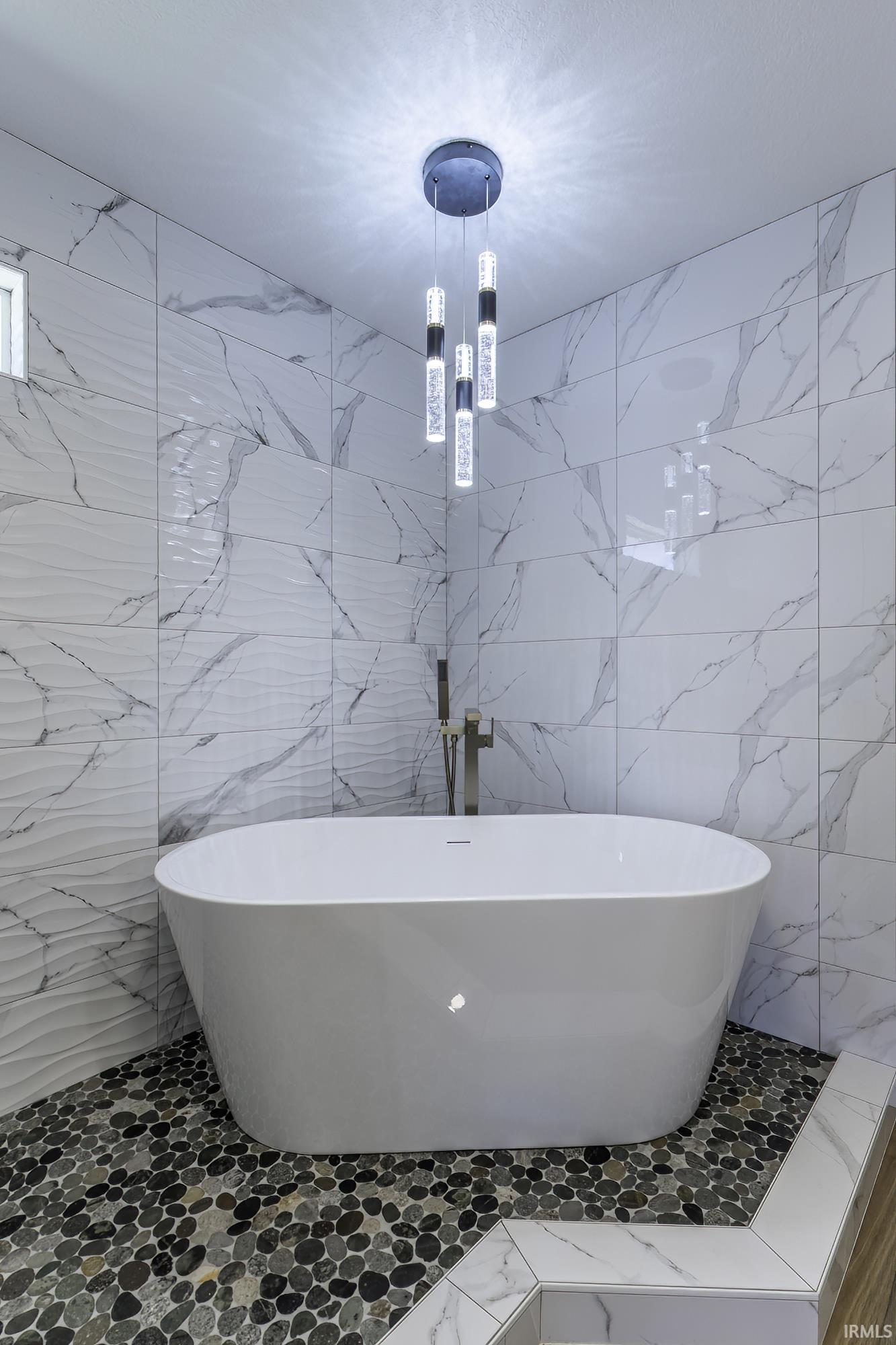 Freestanding soaking tub with a floor-mounted faucet, surrounded by marble-style wall tiling and a pebble-stone floor