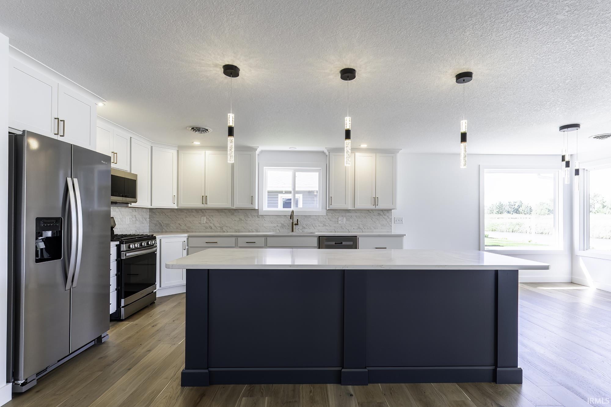 Open-concept kitchen featuring a spacious central island with a light-toned countertop, white cabinetry with contemporary hardware, stainless steel appliances, modern pendant lighting, and wood-finish flooring
