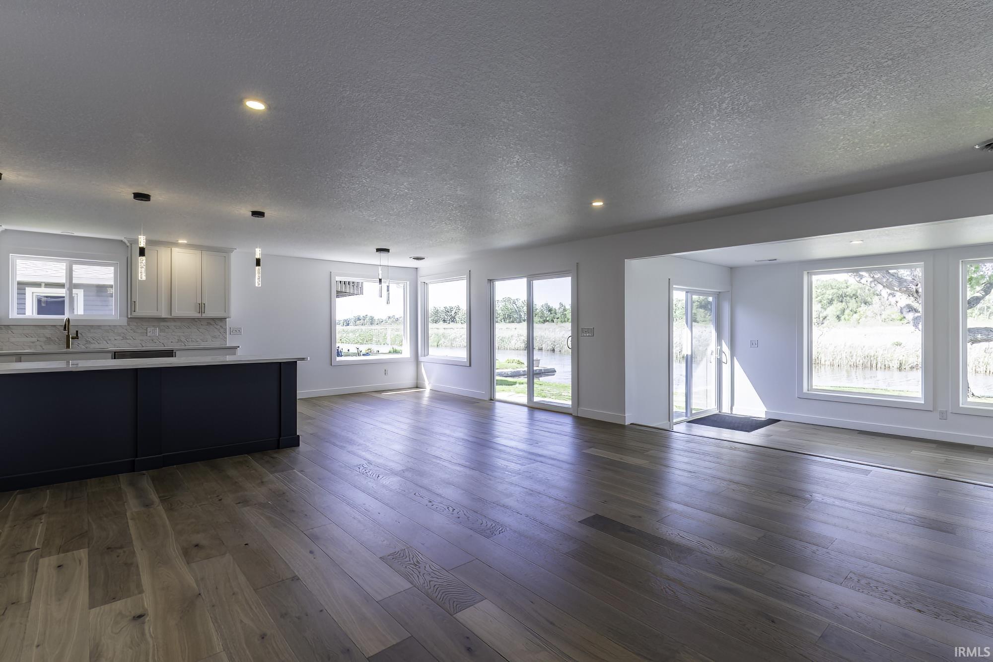 Open-concept living area with wood-finish flooring, a kitchen island with dark cabinetry, white perimeter cabinetry, multiple pendant lights, and numerous windows and glass doors offering abundant natural light