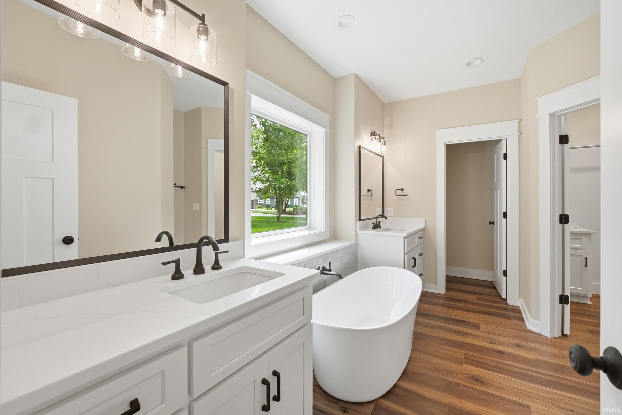 Bathroom featuring a freestanding tub, dual vanity with white cabinetry and countertops, wood-finish flooring, and a large window with a tiled sill