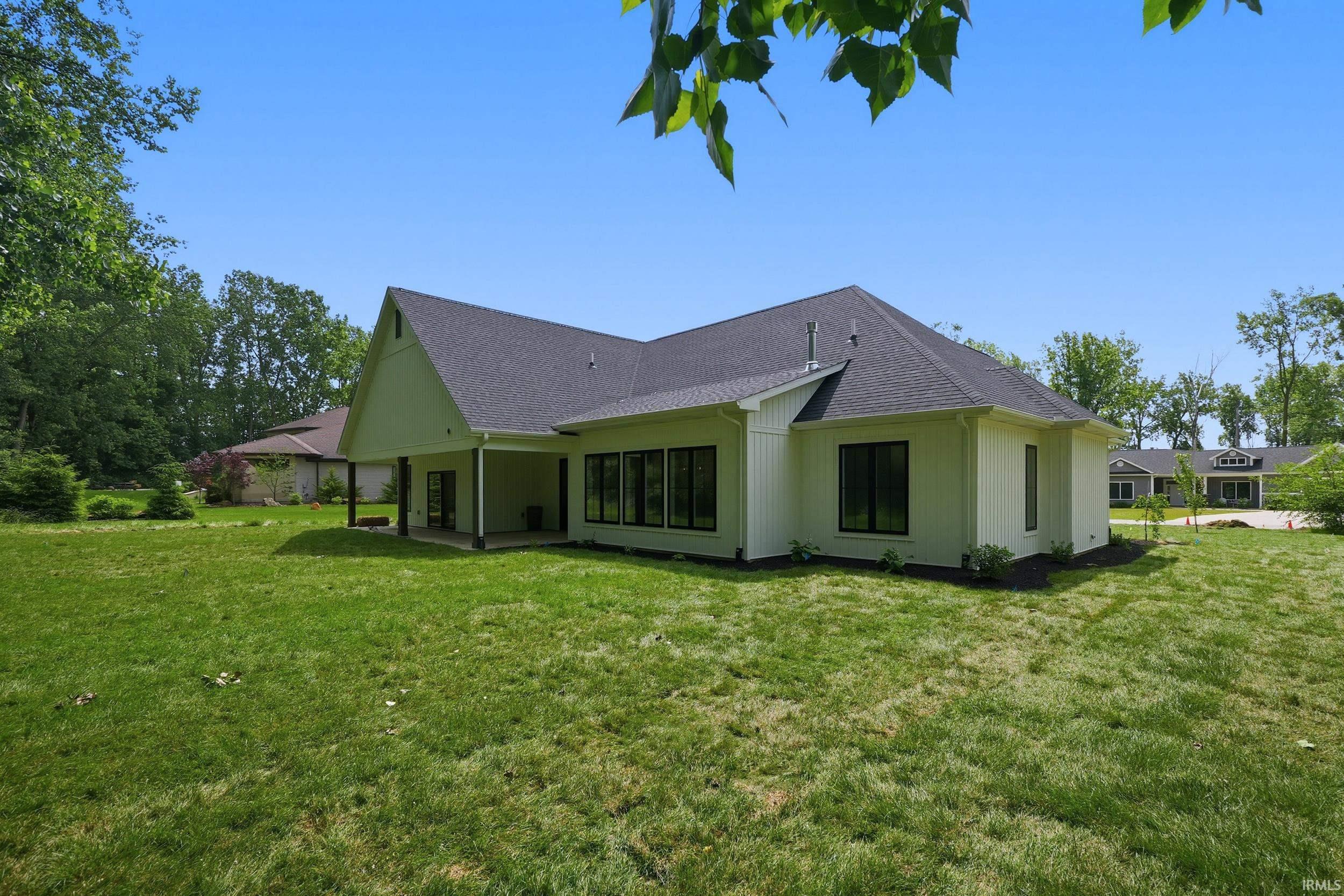 Expansive green lawn surrounds a modern residence featuring a covered patio, numerous large windows with dark frames, light-colored vertical siding, and a dark shingle roof