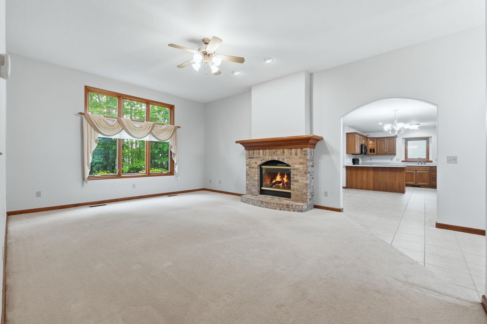 Spacious living area featuring a brick fireplace with a wood mantel, light-toned carpeting, and a multi-pane window with wood-finish trim