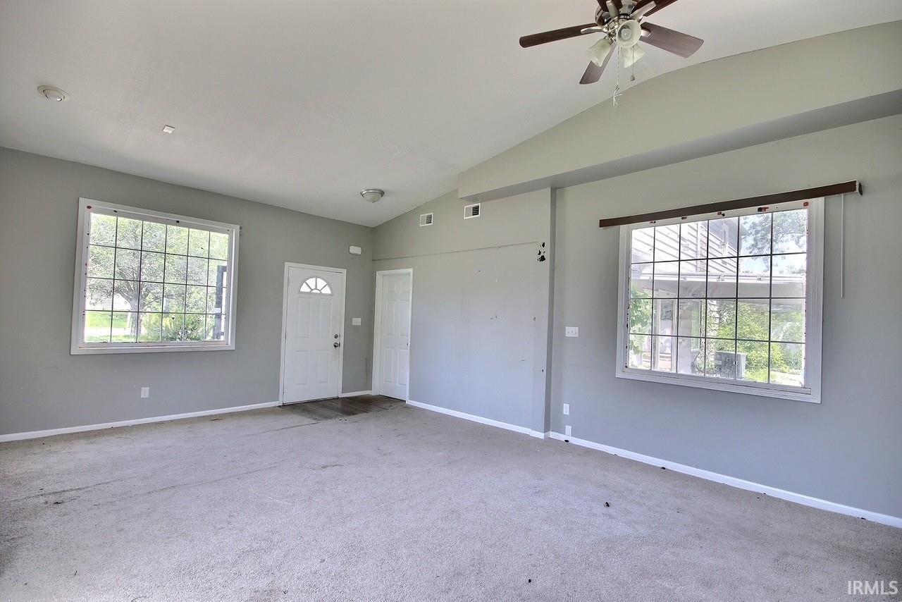 Spacious room featuring vaulted ceilings, a ceiling fan, two large windows with grid patterns, and a white paneled entry door