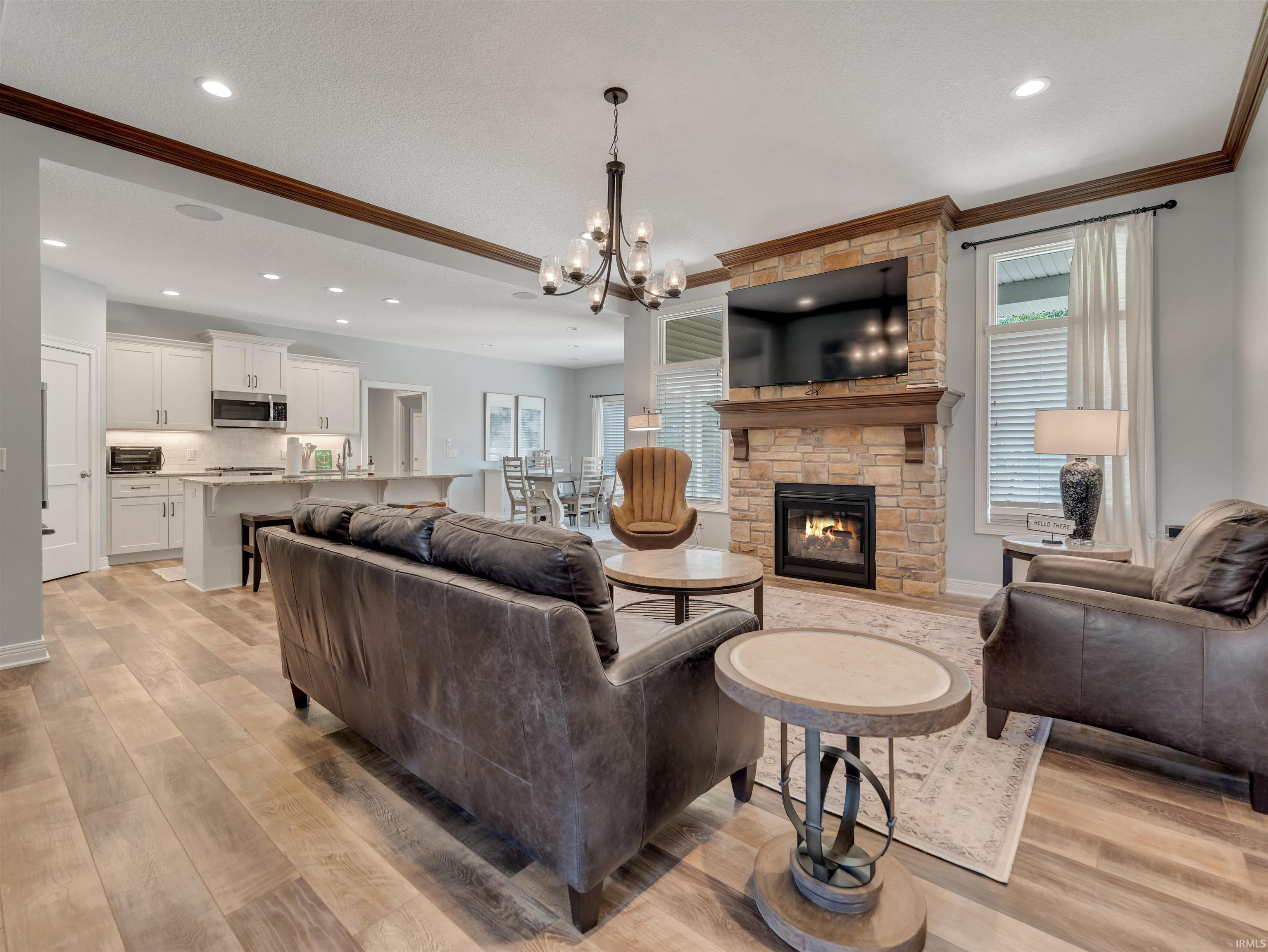 Open-concept living space featuring wood-finish flooring, a stone-surround fireplace with a wood mantel, a chandelier, recessed lighting, and white kitchen cabinetry with stainless steel appliances