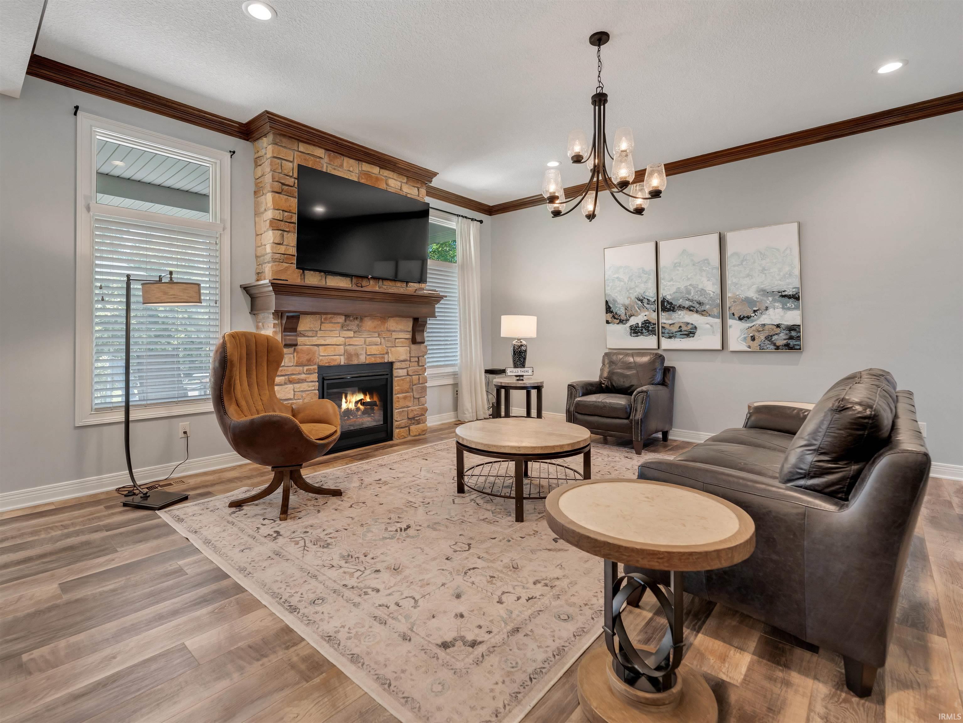 Spacious living area featuring a stone-clad fireplace with wood mantle, wood-finish flooring, crown molding, and a multi-arm chandelier