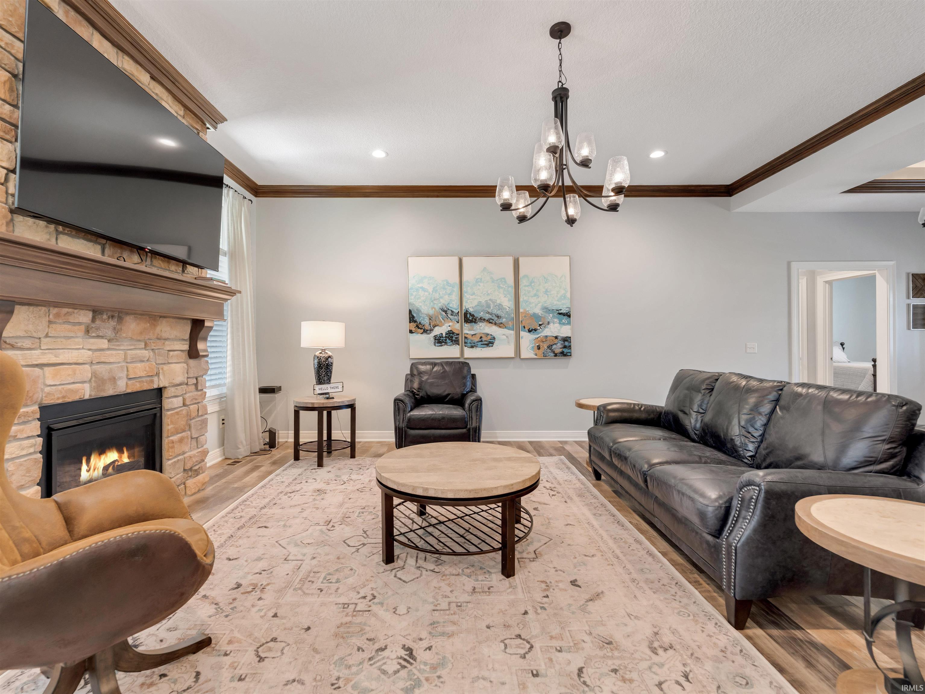 Living space featuring a stacked stone fireplace with a wood mantle, wood-finish flooring, recessed lighting, crown molding, and a central chandelier