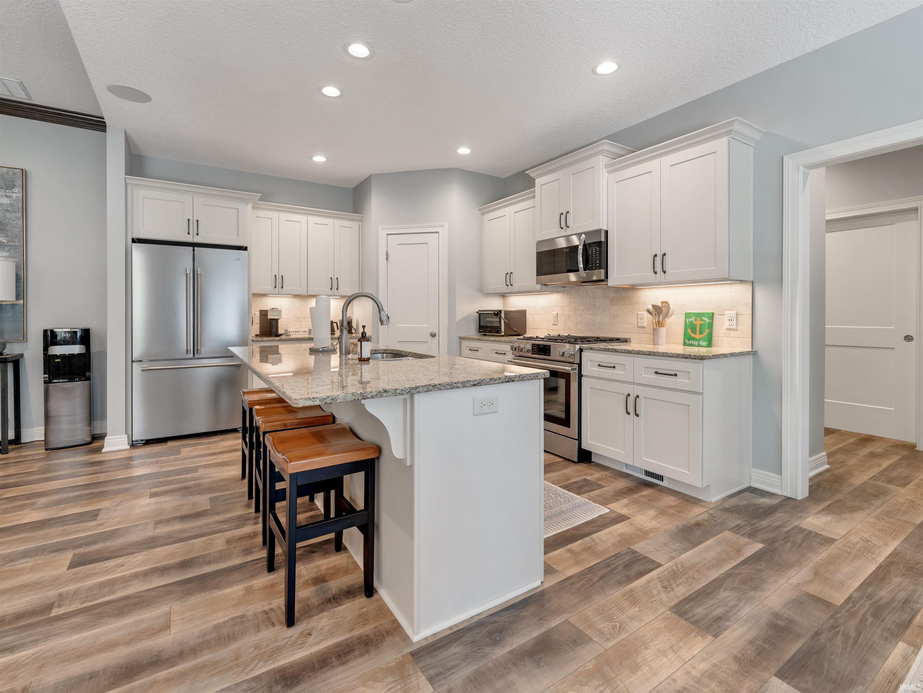 Spacious kitchen featuring an expansive center island with seating, stainless steel appliances, white shaker cabinetry, granite countertops, and wood-finish flooring
