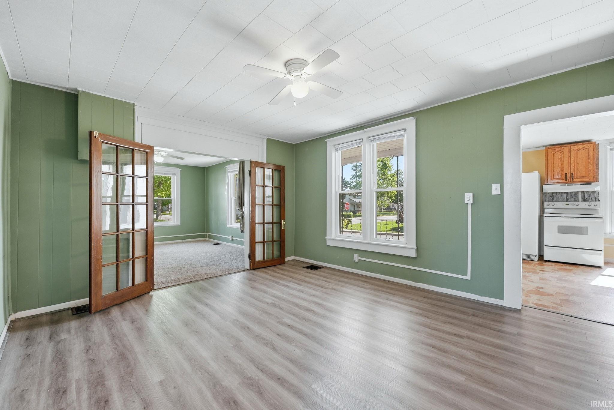 Spacious room featuring wood-finish flooring, two large windows with white trim, a ceiling fan, and French doors leading to an additional room