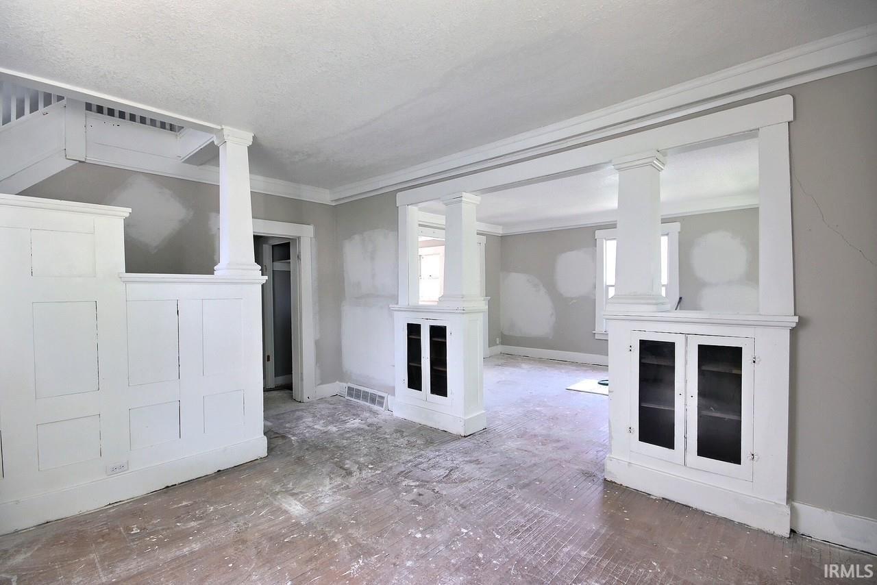 Interior space featuring wood-finish flooring, half-wall paneling, built-in cabinetry with glass doors, and decorative columns