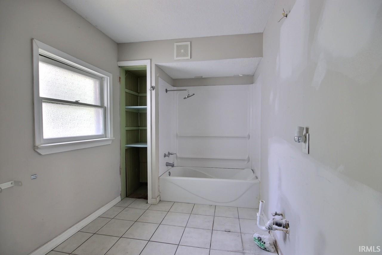 Bathroom featuring a white bathtub/shower combination, built-in shelving, white tiled flooring, and a window with frosted glass