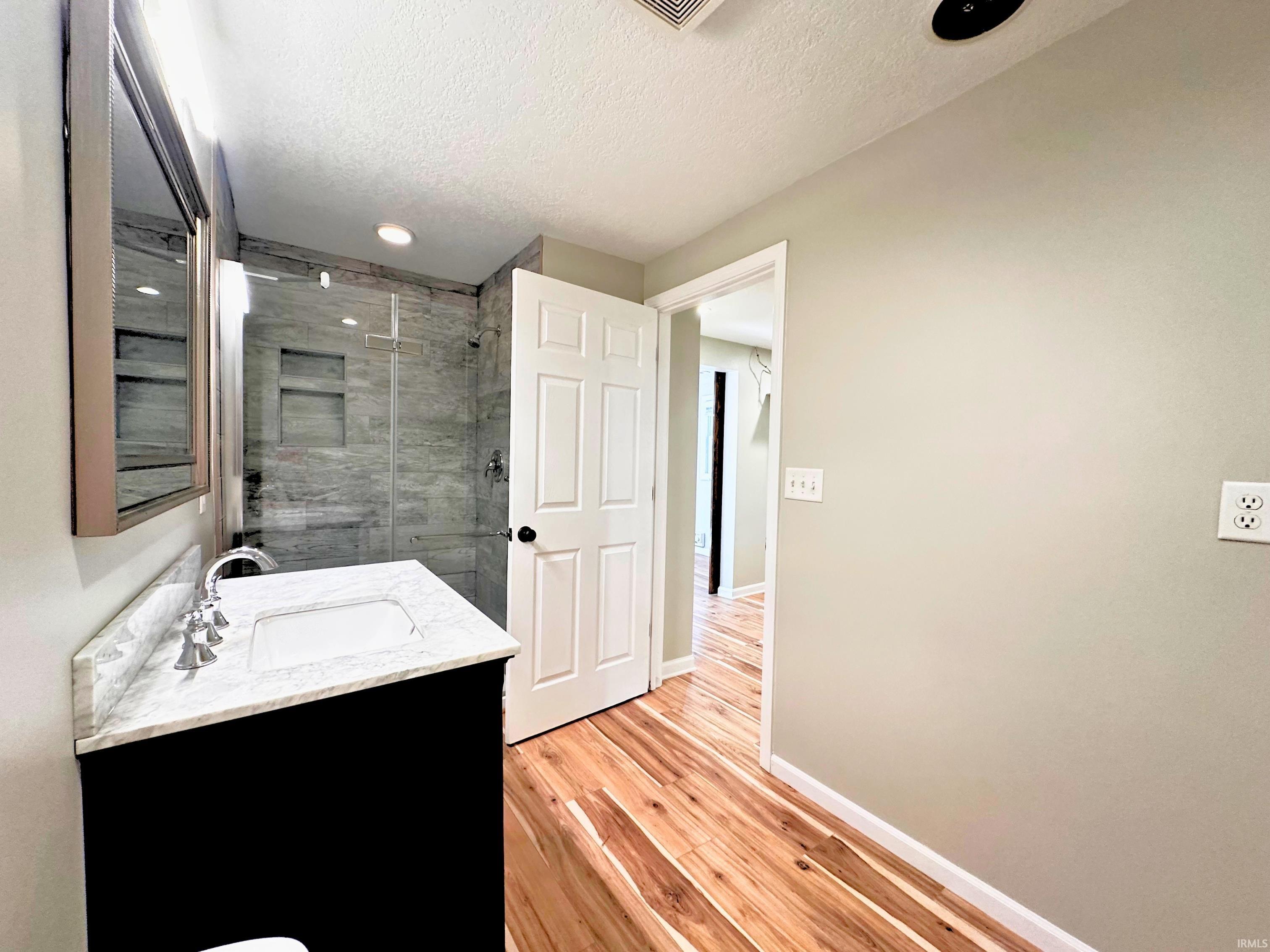 Bathroom featuring a dark wood vanity with a white countertop and undermount sink, a large mirrored medicine cabinet, a glass-enclosed shower with gray tiled walls and built-in niches, and wood-finish flooring