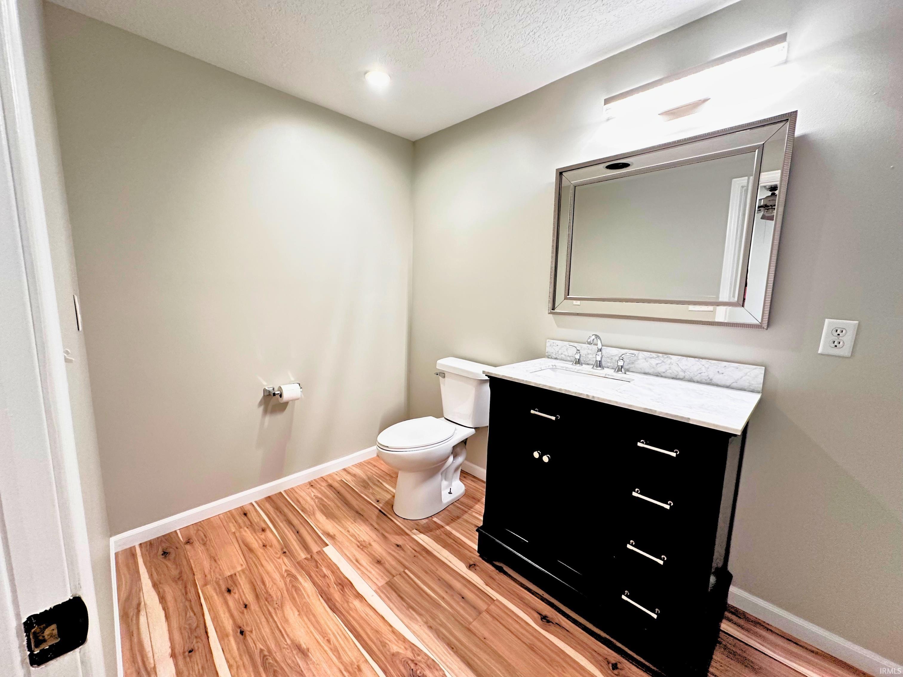 Bathroom featuring wood-finish flooring, a contemporary black vanity with a white countertop, a rectangular mirror with a silver-toned frame, and overhead vanity lighting