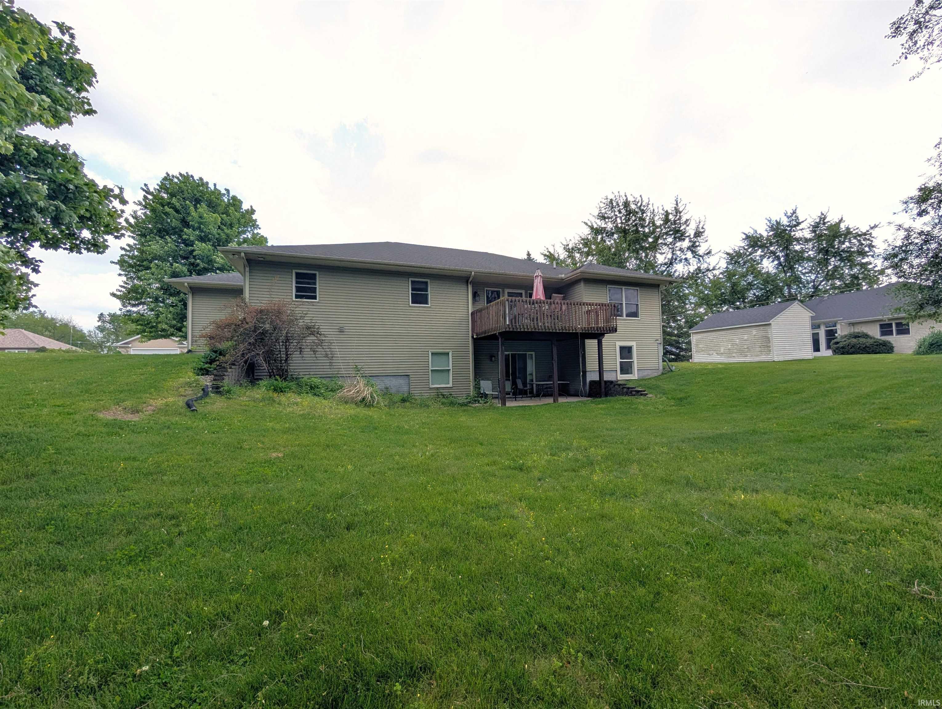 Expansive green lawn with a multi-level house featuring a second-story wooden deck, ground-level patio, and a light olive-toned exterior siding