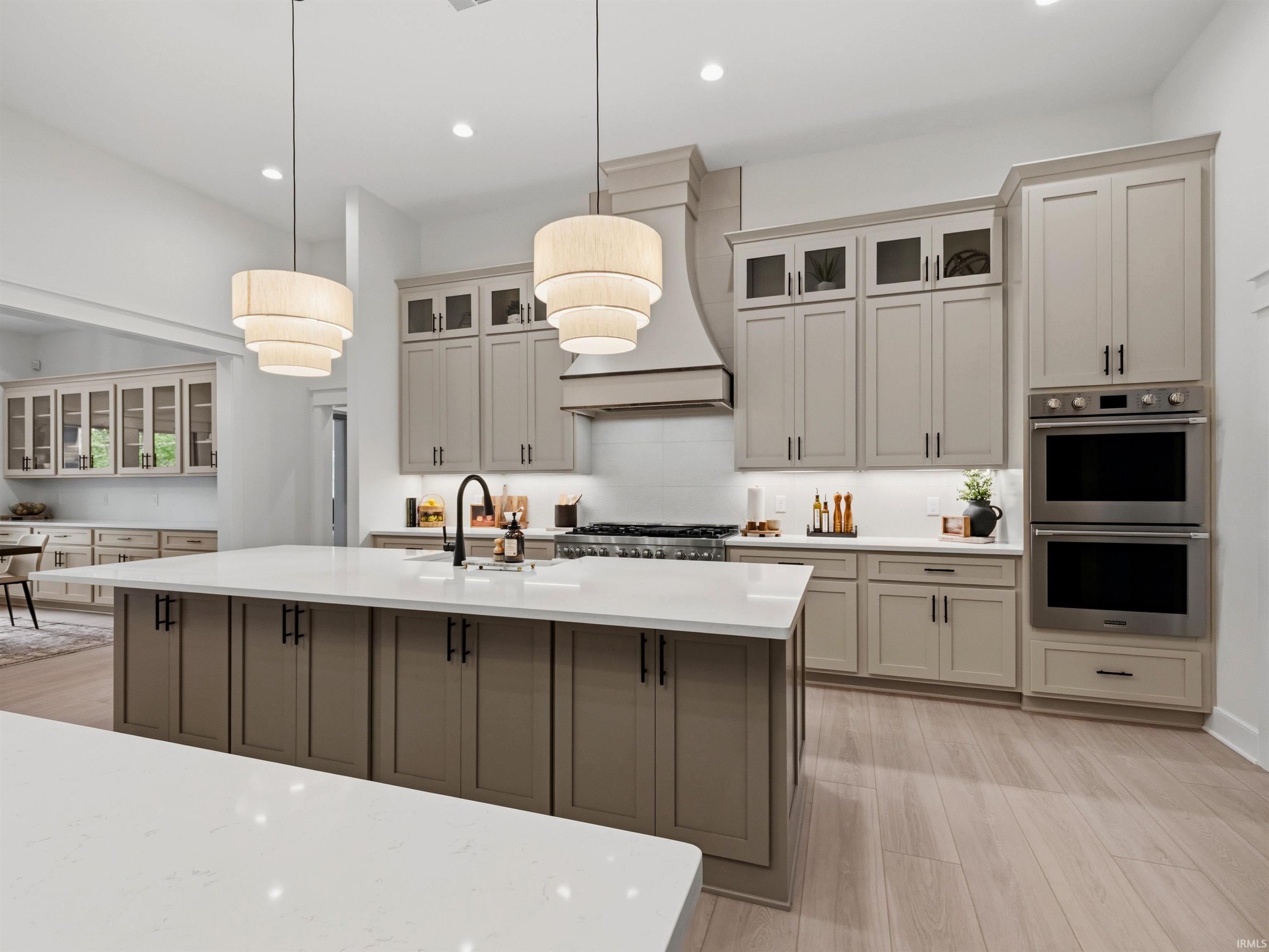Kitchen featuring a large central island with a white countertop and dark base cabinetry, light wood-finish flooring, two tiered pendant lights, a built-in double oven, and recessed lighting