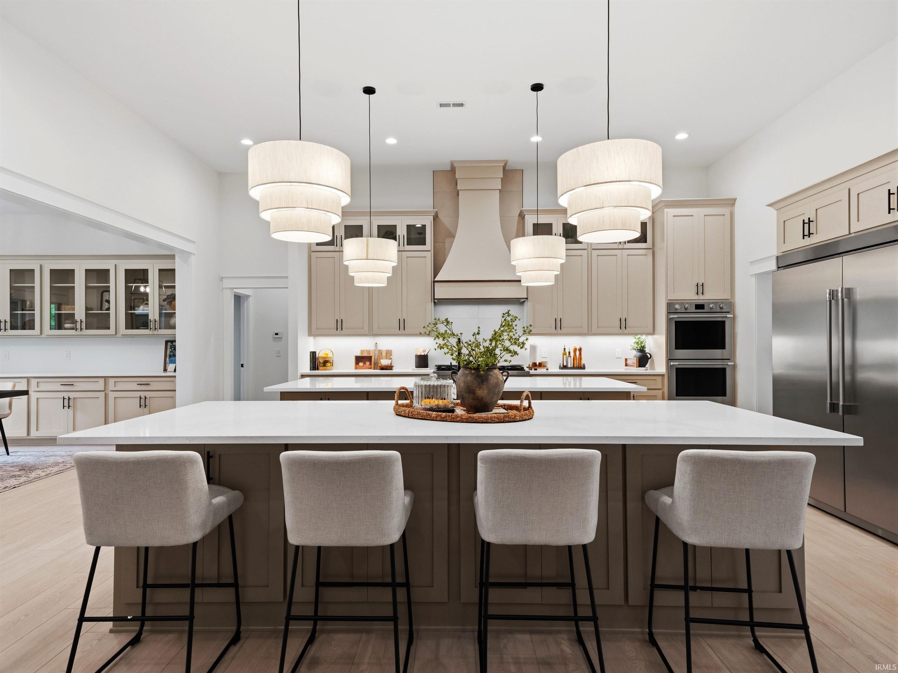 Kitchen featuring dual islands with white countertops, light wood-finish flooring, custom cabinetry with integrated appliances, a decorative range hood, and tiered pendant lighting