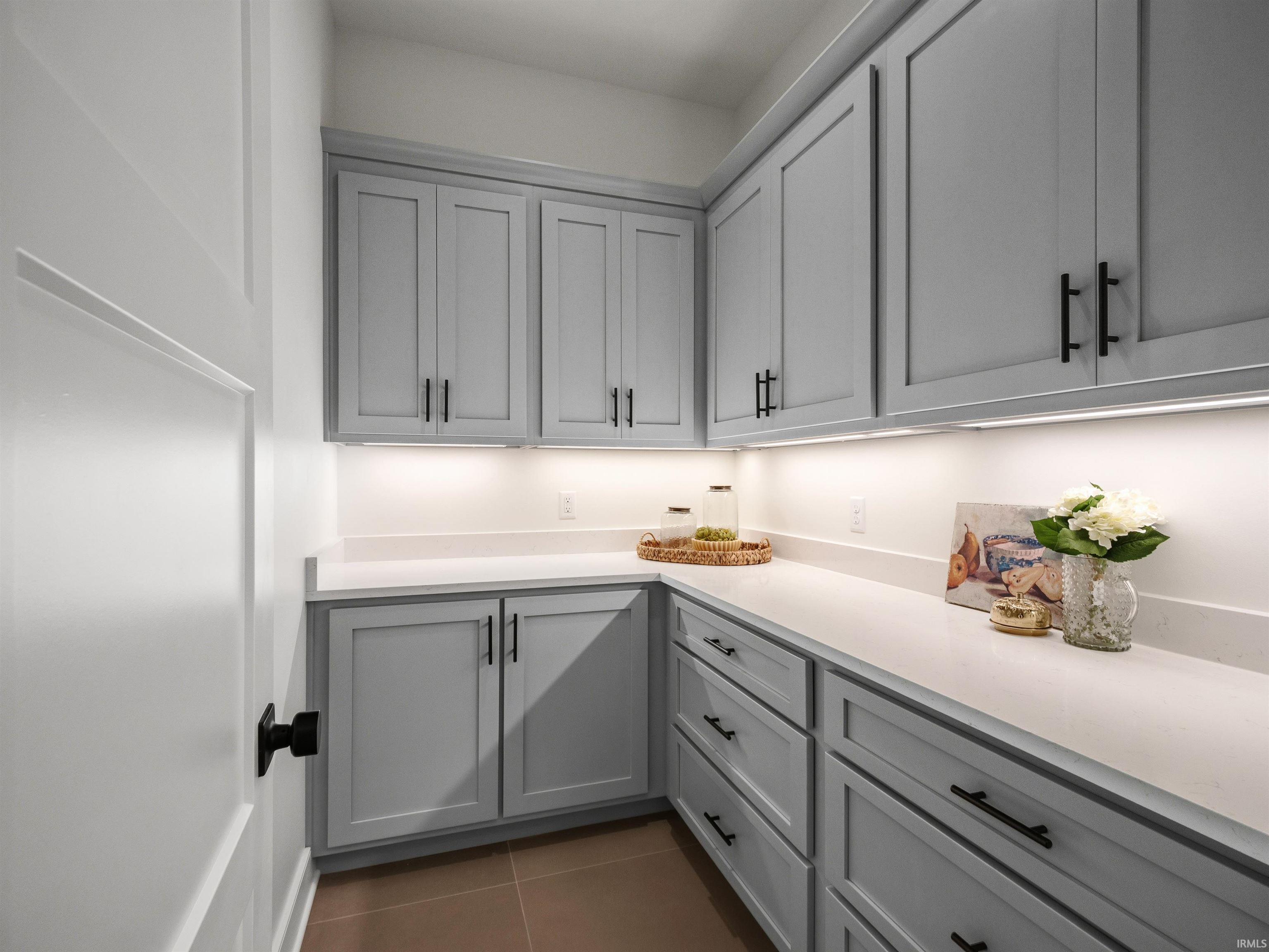 Kitchen pantry featuring extensive gray shaker-style cabinetry with matte black hardware, under-cabinet lighting, light-toned countertops, and tiled flooring