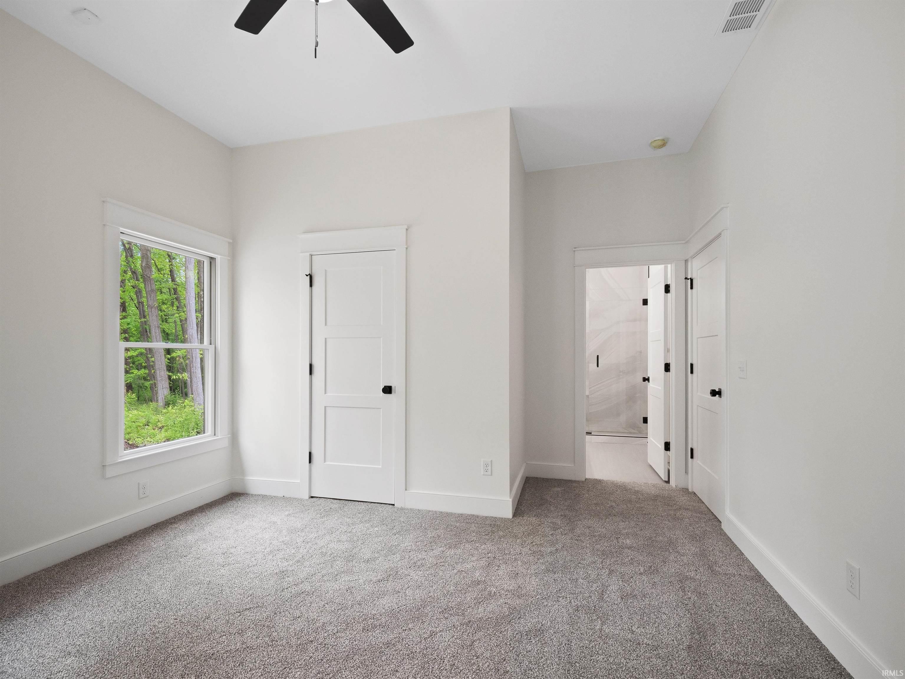 Neutral-toned room featuring a single window with a wooded backdrop, plush gray carpeting, a contemporary black ceiling fan, and white doors with matte black hardware