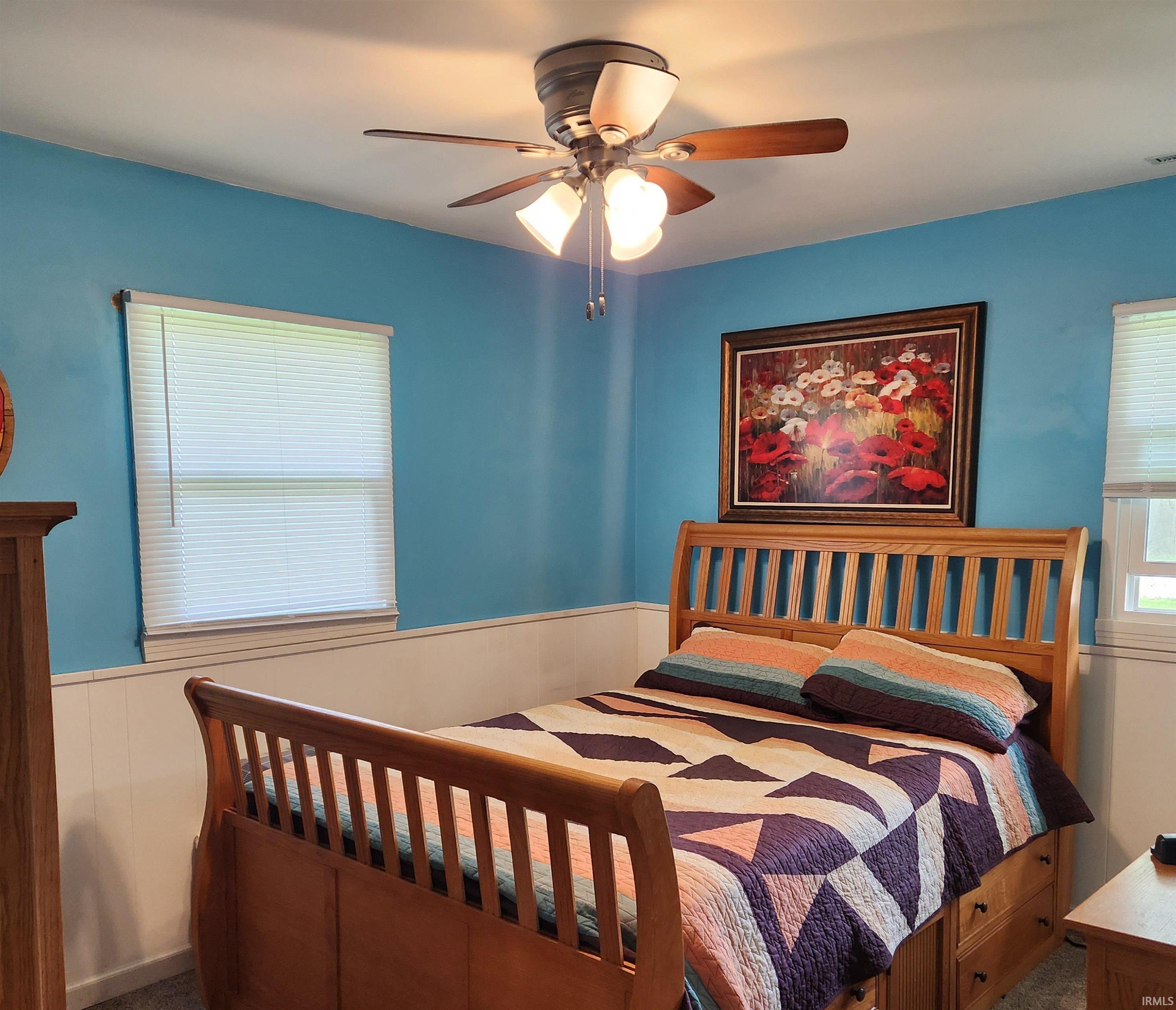 Bedroom featuring paneling wainscoting, two windows with blinds, a ceiling fan with integrated lighting, and carpet flooring