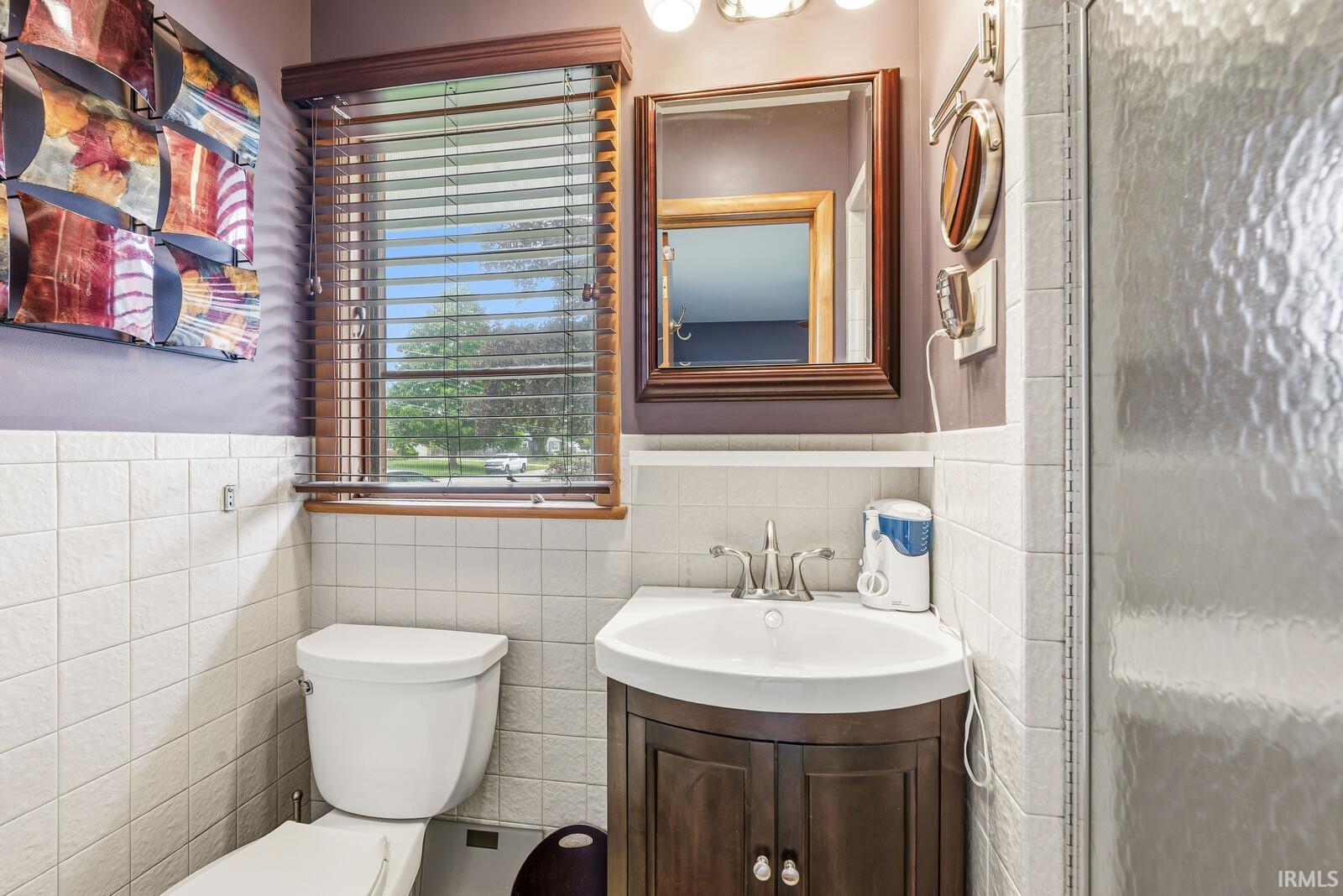 Bathroom featuring white tile walls, a corner vanity with a wood-finish cabinet and integrated sink, and a toilet