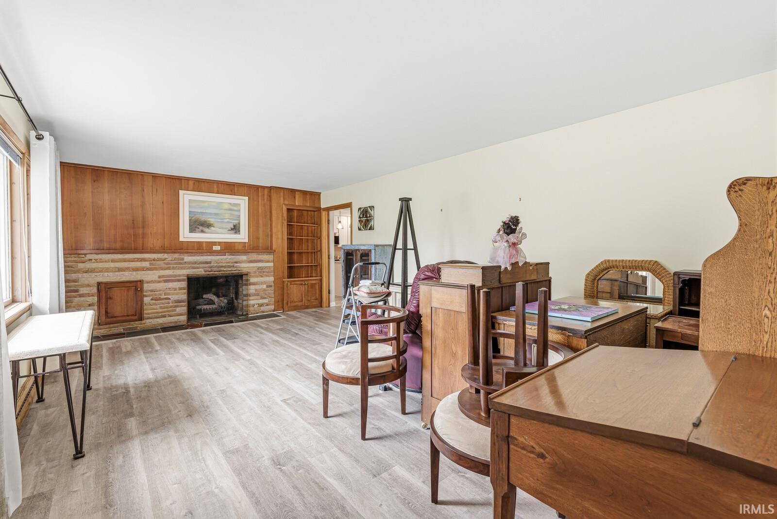 Living area featuring wood-finish flooring, a stone-faced fireplace, a wood paneled accent wall, a built-in bookshelf, and large windows with white curtains