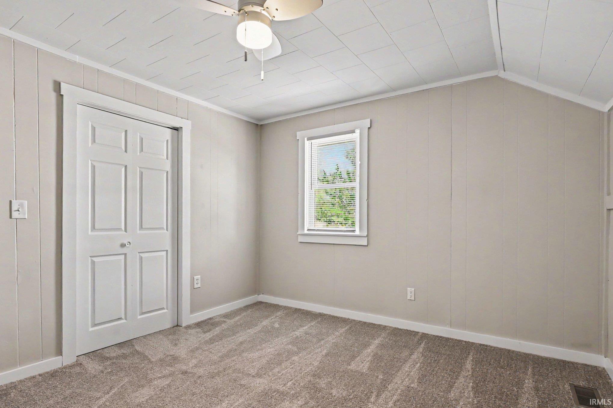 Carpeted room featuring a ceiling fan, a window with exterior tree scenery, a paneled accent wall, and bi-fold closet doors