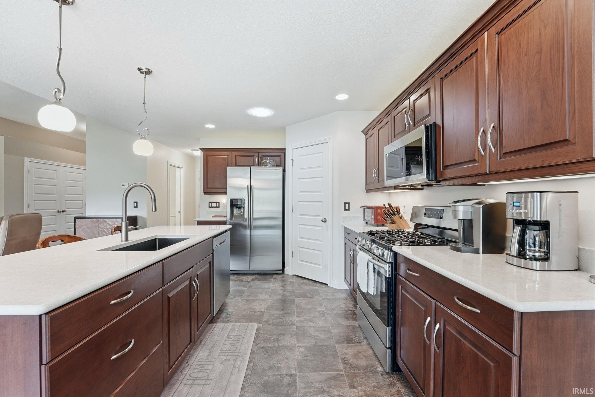 Modern kitchen featuring an expansive center island with an undermount sink, stainless steel appliances, abundant wood cabinetry, light-toned countertops, and durable tile flooring