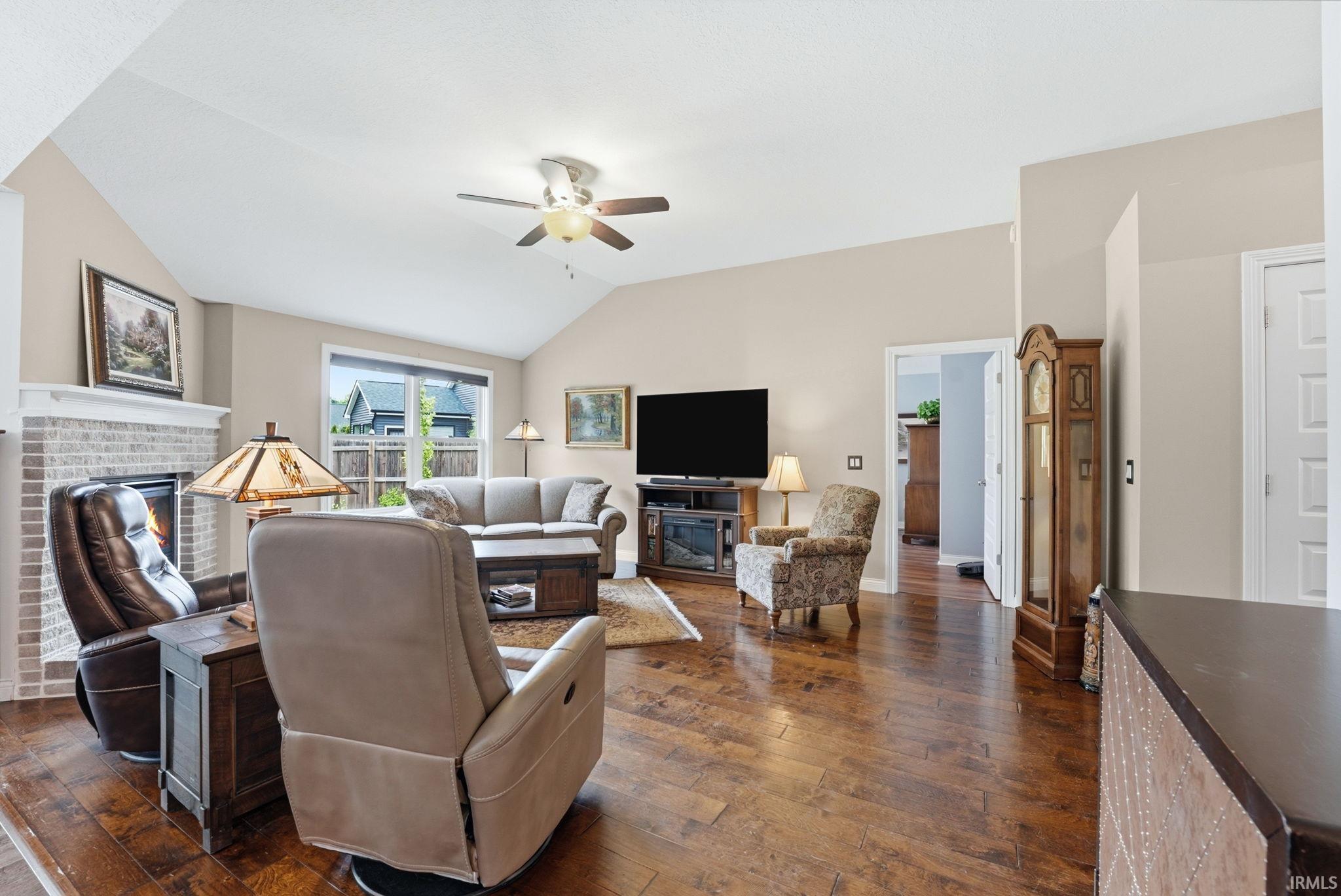 Living room featuring vaulted ceilings, wood-finish flooring, a brick fireplace with mantel, and a ceiling fan