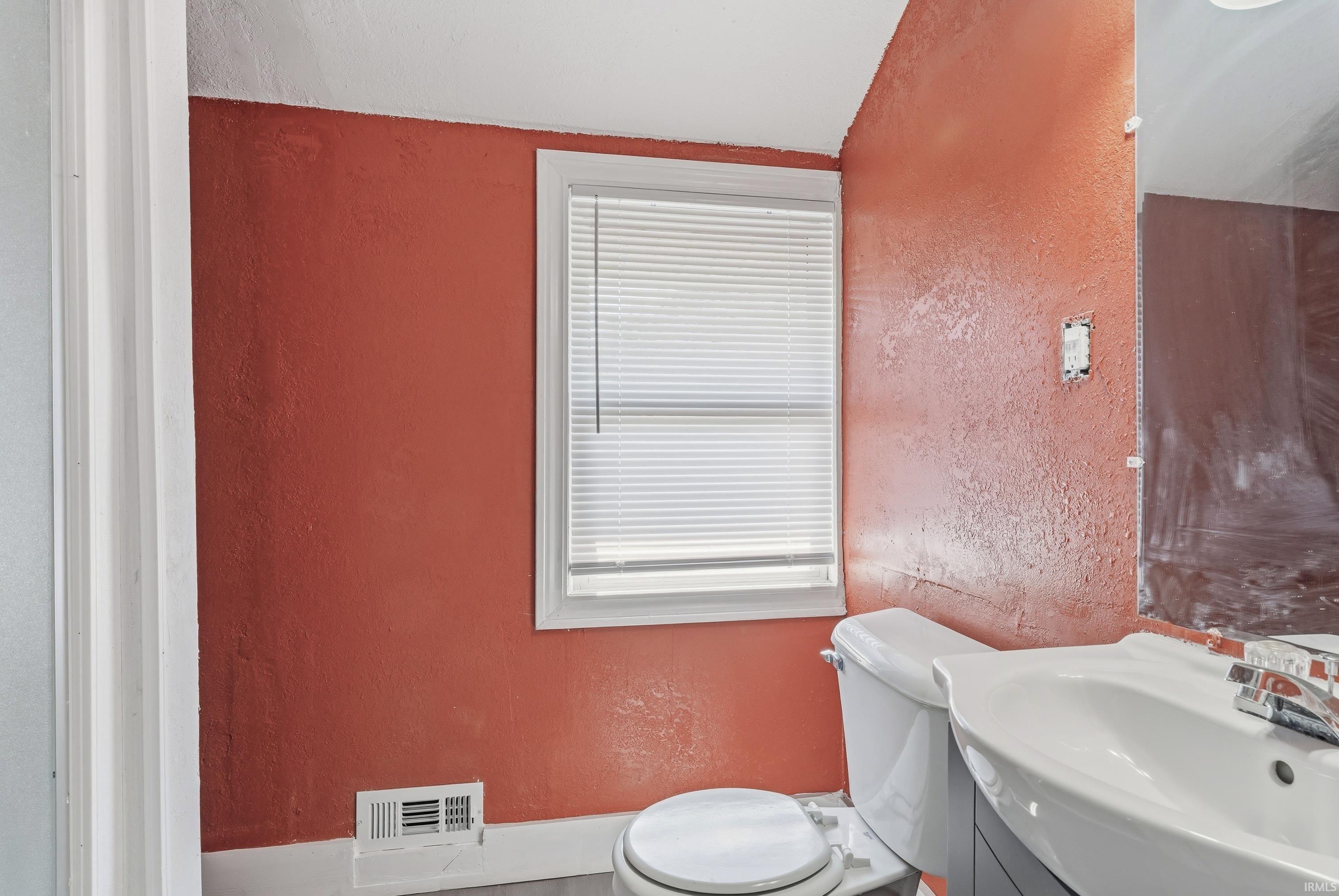Bathroom featuring a white window with blinds, a wall-mounted sink with a chrome faucet, a white toilet, and gray wood-finish flooring