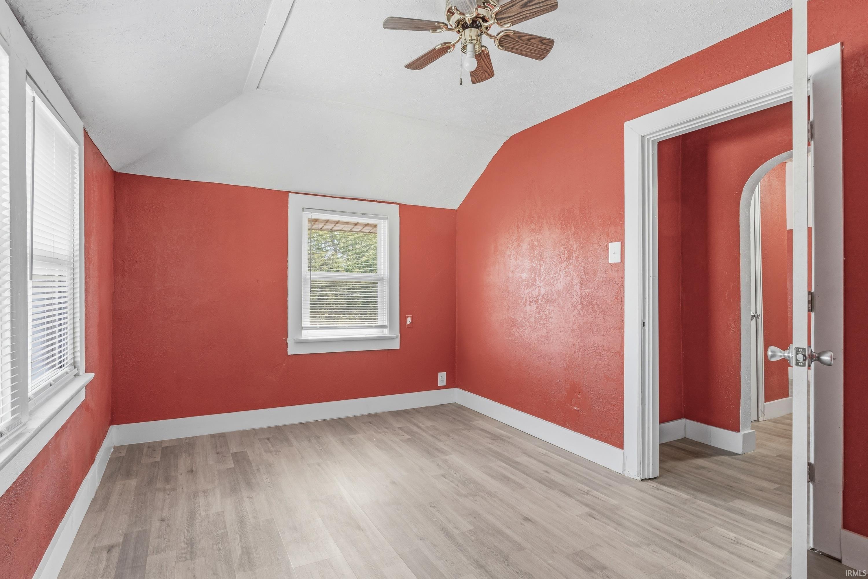 Attic room featuring wood-finish flooring, two windows with blinds, a ceiling fan, and white trim