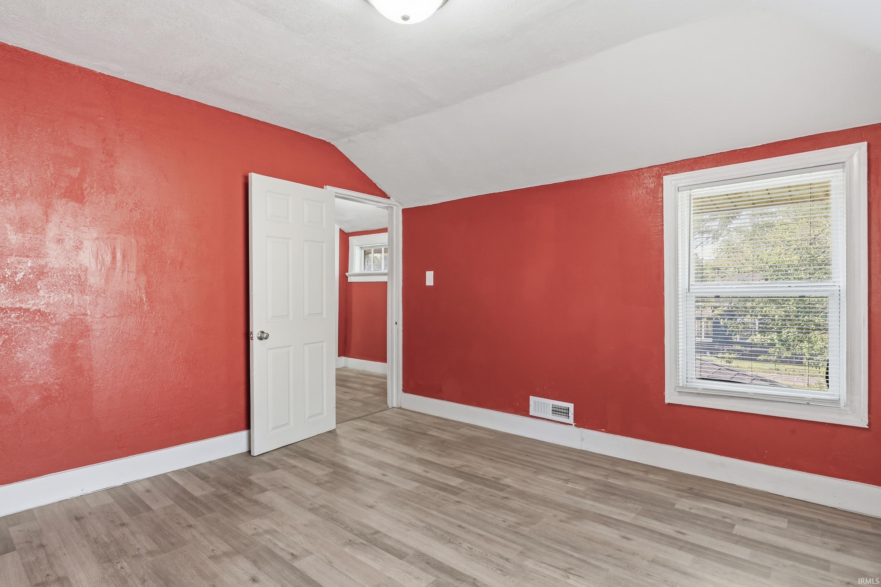 Spacious room featuring wood-finish flooring, vibrant red walls, white baseboards, and a window with blinds
