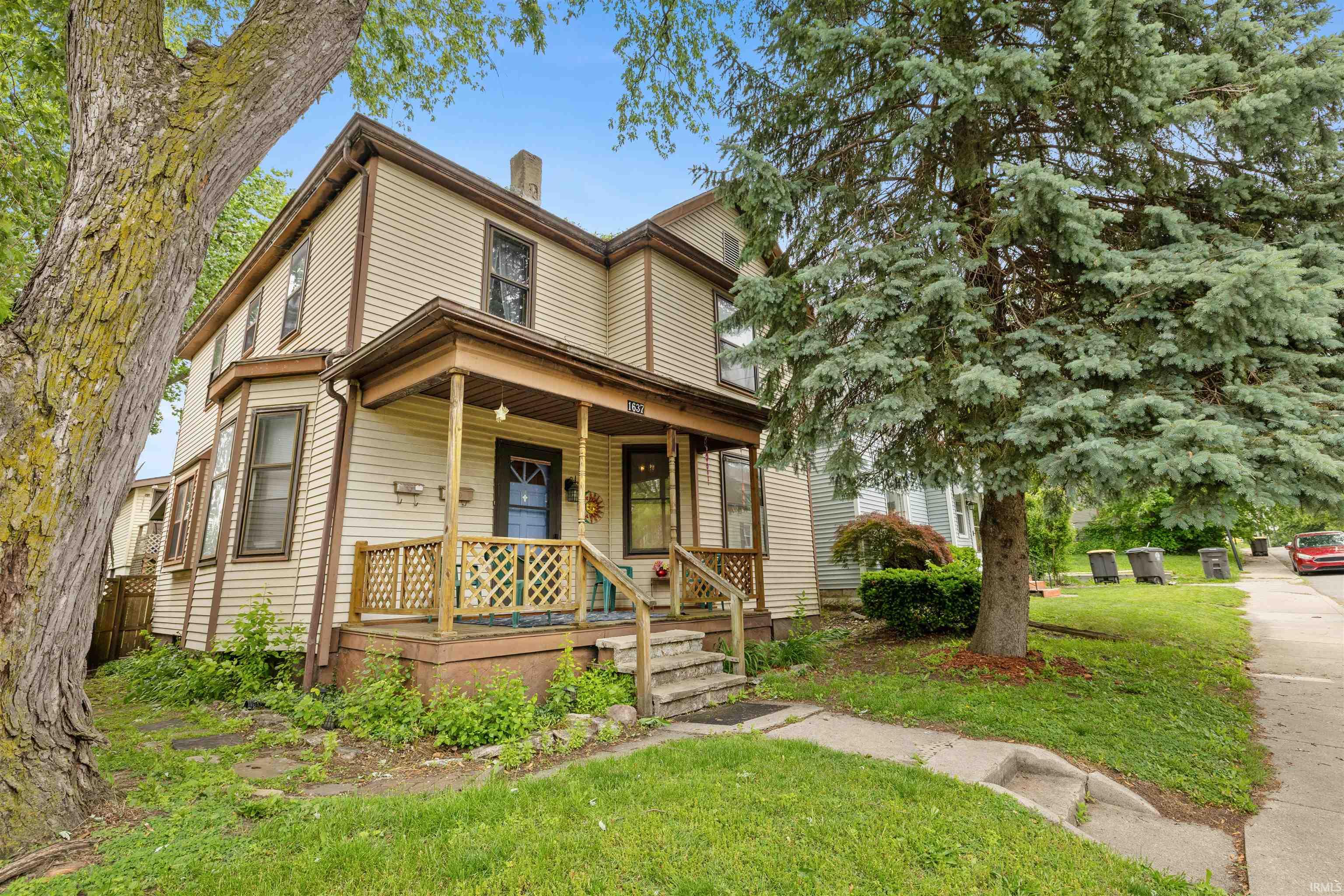 Two-story residence featuring light-colored siding, a covered front porch with lattice railing, bay windows, and a prominent evergreen tree in the front yard