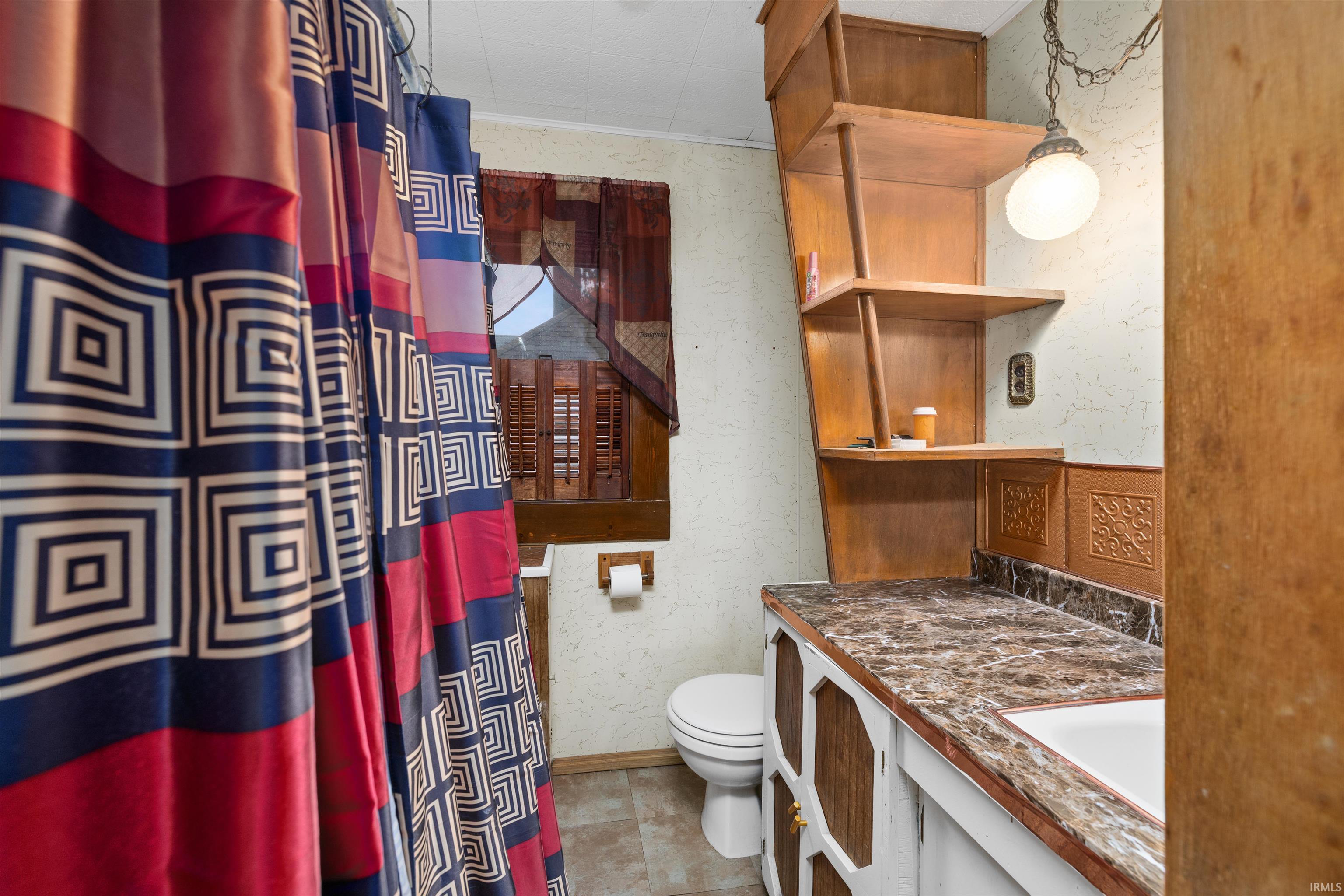 Bathroom featuring a built-in wood shelving unit, vanity with a rectangular sink, patterned countertop, and wood-paneled window with blinds