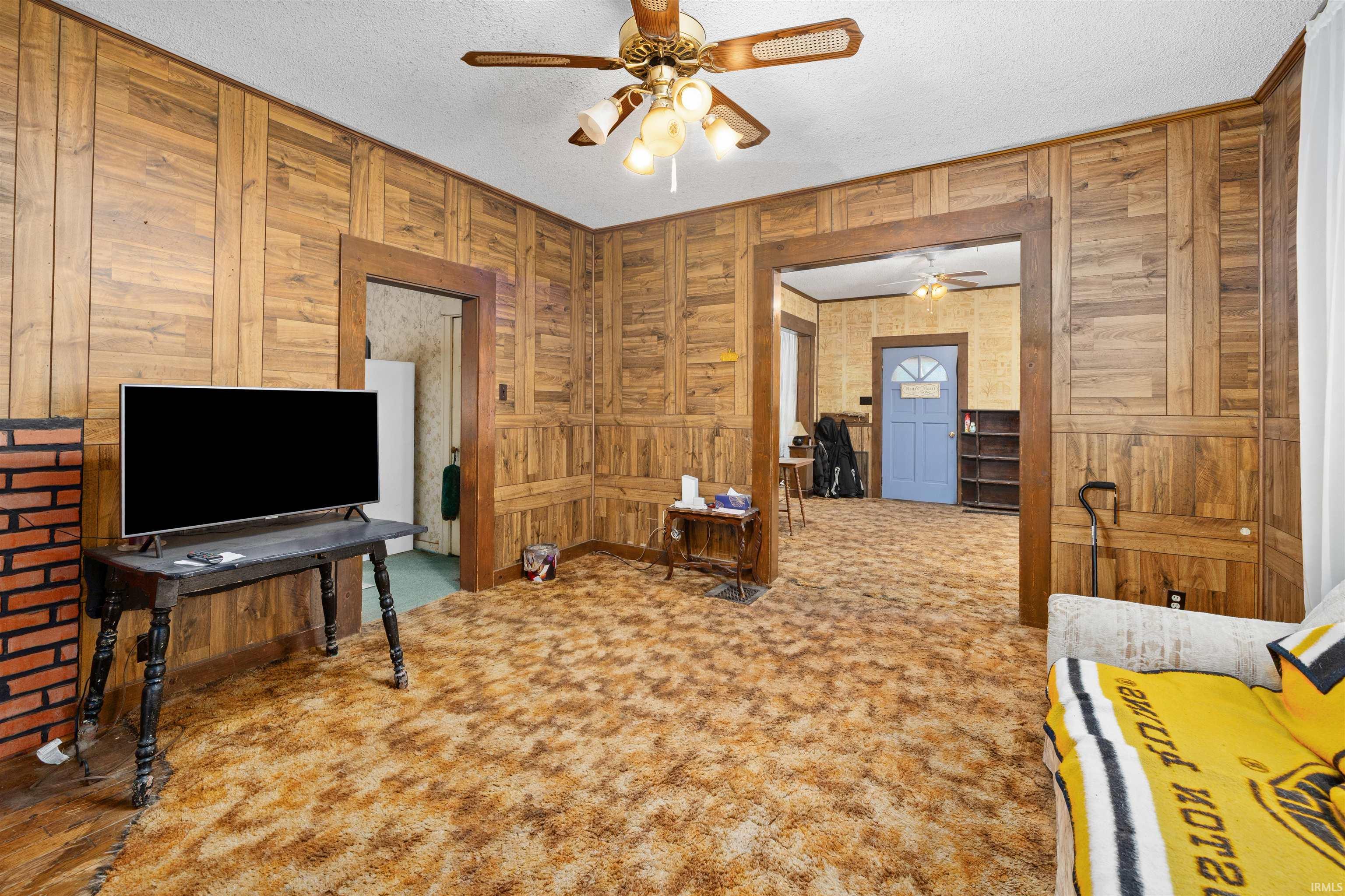Living area featuring wood paneling throughout, a brick-faced fireplace, and a ceiling fan with integrated lighting