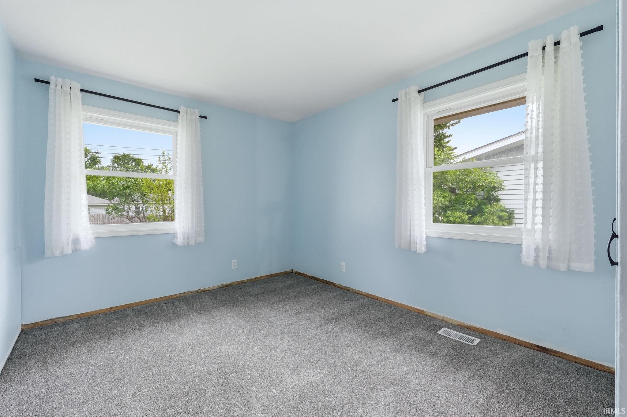 Carpeted room featuring two windows with white trim, light blue walls, and a visible HVAC register