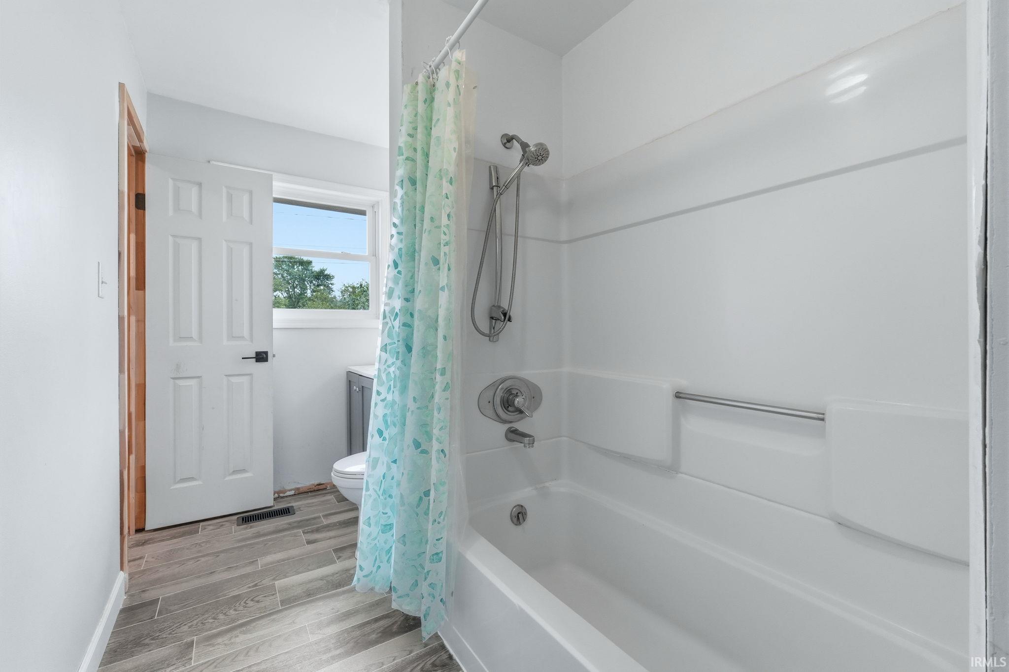 White bathtub with a chrome shower fixture, light gray wood-finish flooring, white paneled door, and a window