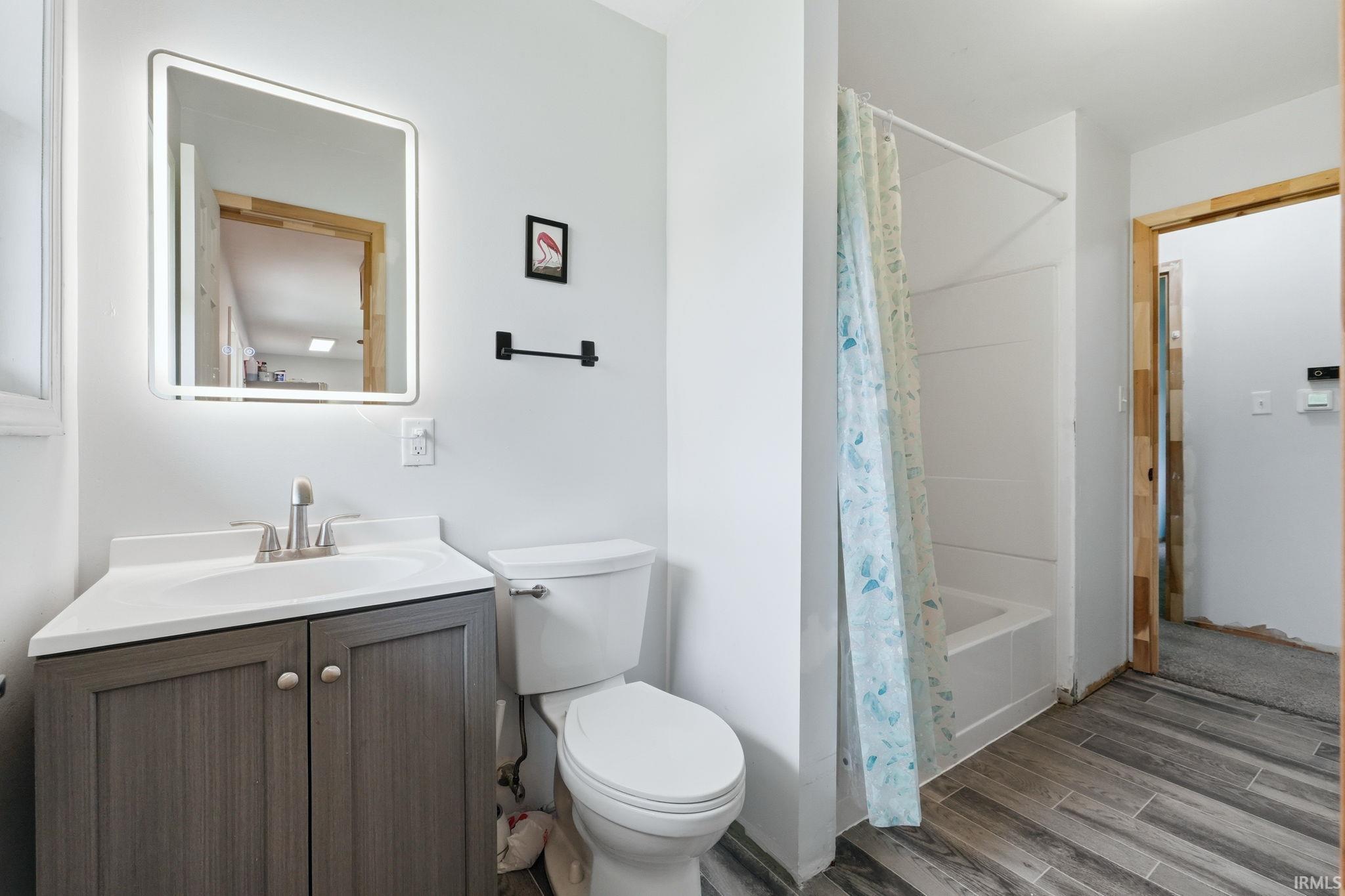 Bathroom featuring a vanity with a white countertop and undermount sink, a backlit mirror, a toilet, a tub/shower combination, and wood-finish flooring