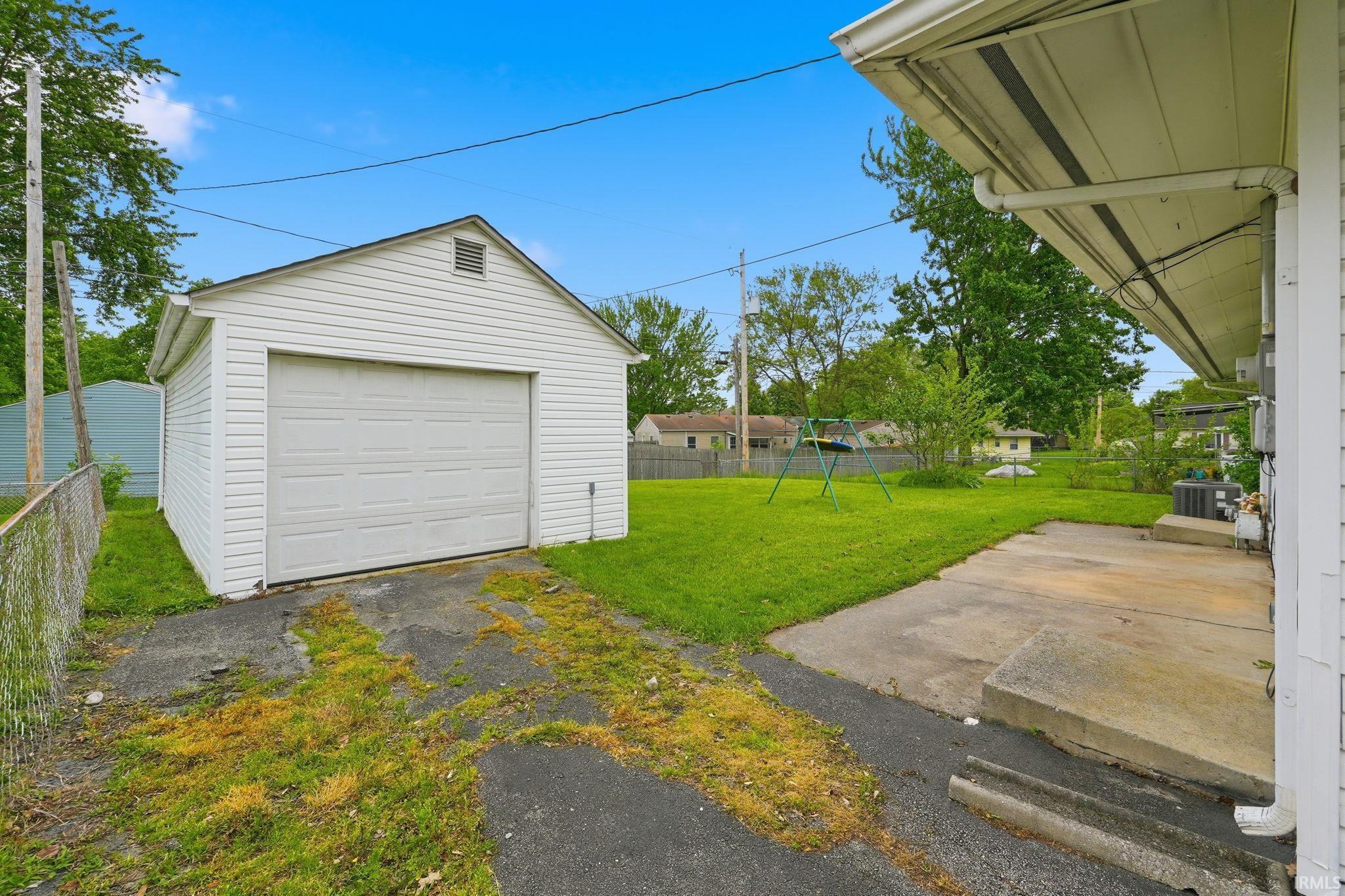 Detached white vinyl-sided garage with a single vehicle door and gable vent, accompanied by an extensive grass yard and a concrete patio
