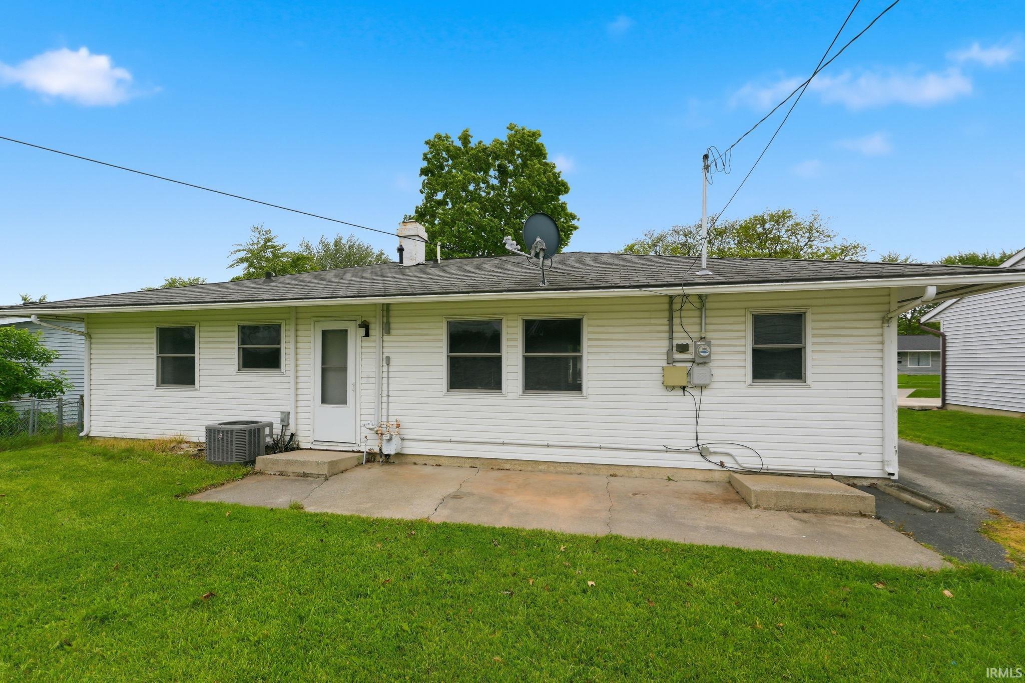 Rear exterior featuring white horizontal siding, a dark shingle roof, multiple windows, and a concrete patio